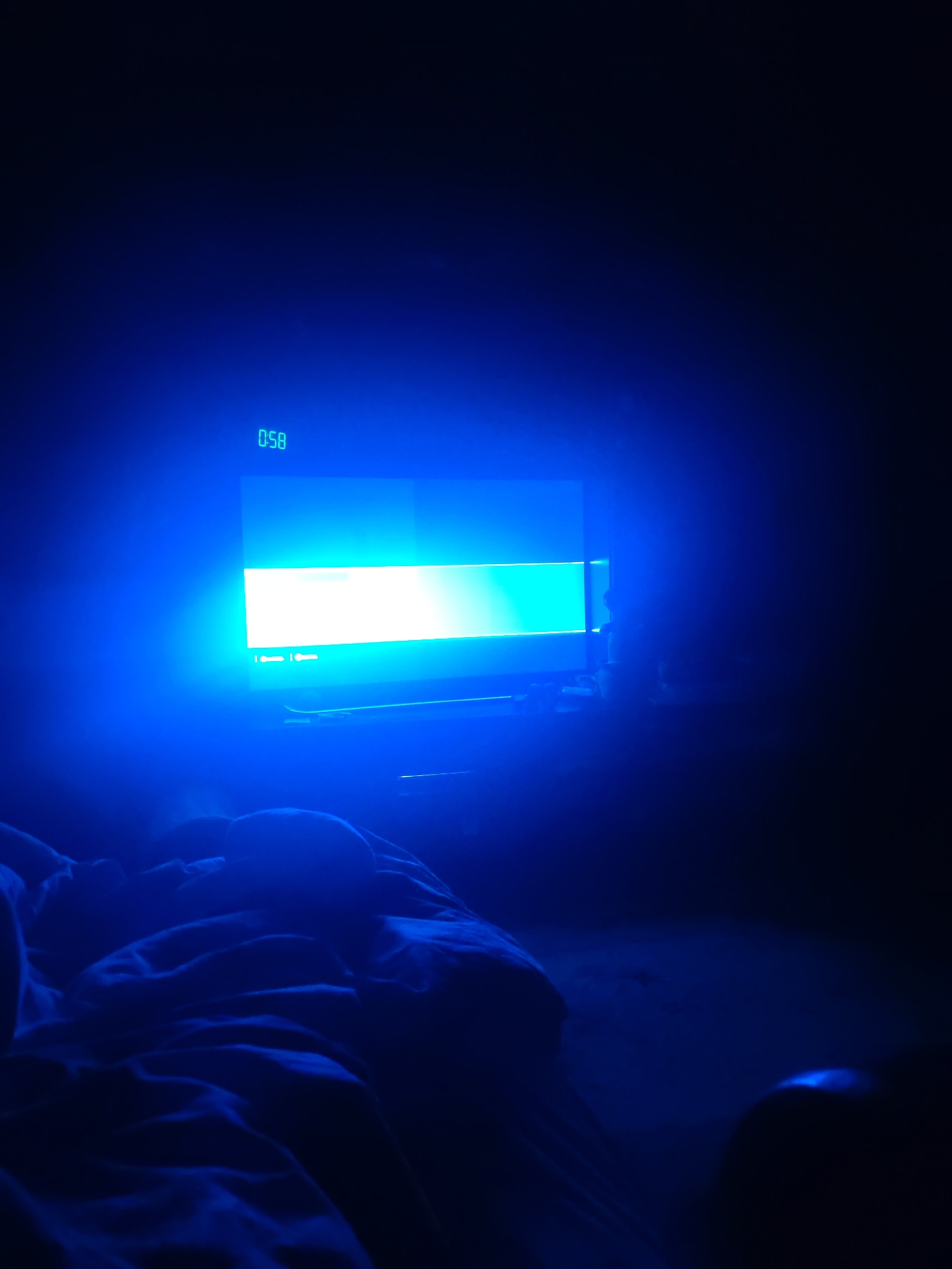 The image shows a dimly lit room with a television screen displaying a bright blue light. A digital timer on the screen reads "0:58". In the foreground, a person is lying in bed, covered by dark blue blankets. The person's head and upper body are visible, and they appear to be asleep or resting. The overall mood of the image is calm and serene. There is no information about the location within the city of Vladimir, Russia.