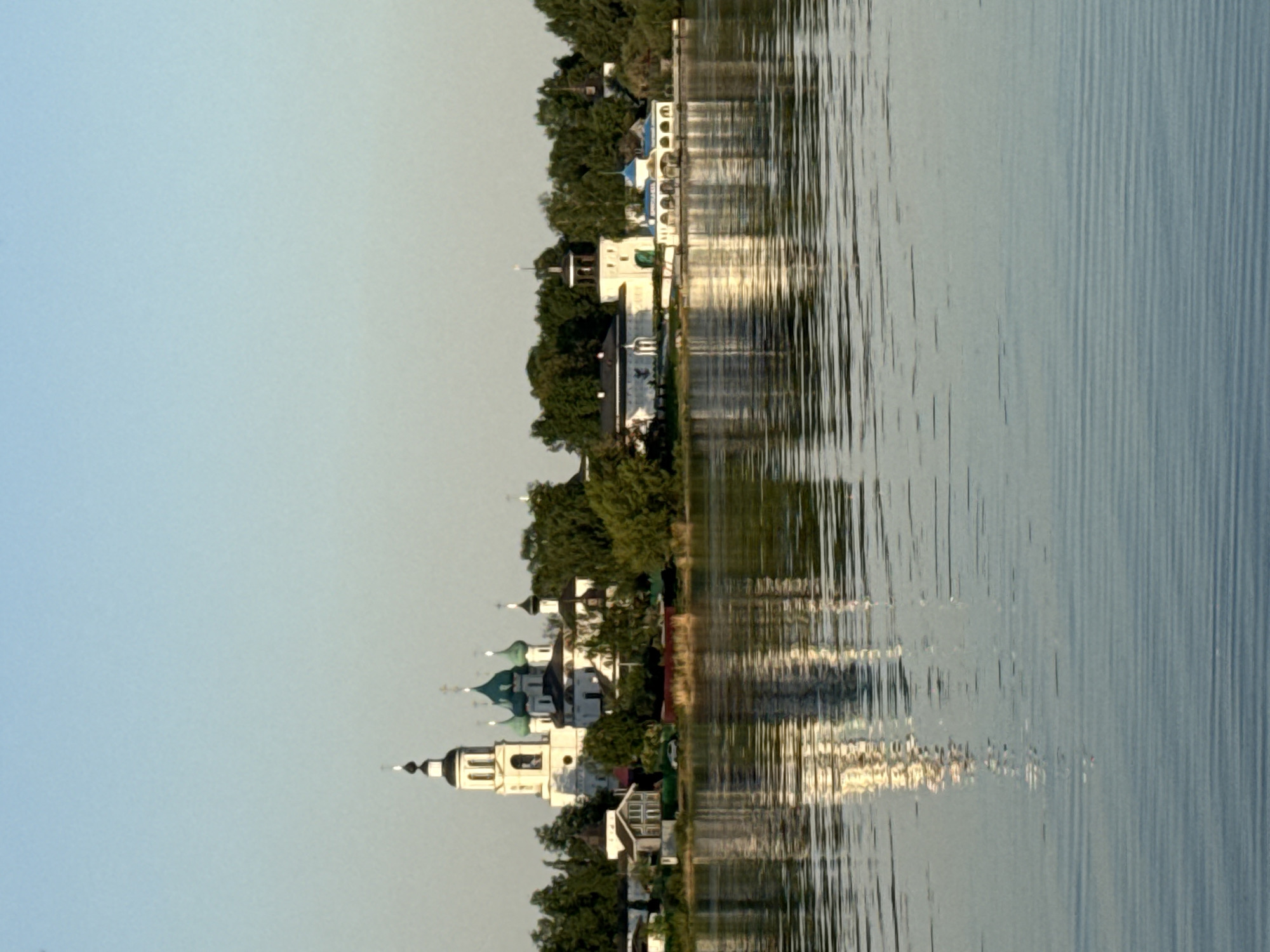 The image depicts a shoreline in Rusanovo, Russia, with buildings and trees reflected in the water. A church with green domes and a bell tower is visible on the shoreline. The water shows vertical striations, likely caused by wind or current. The sky is a light blue.