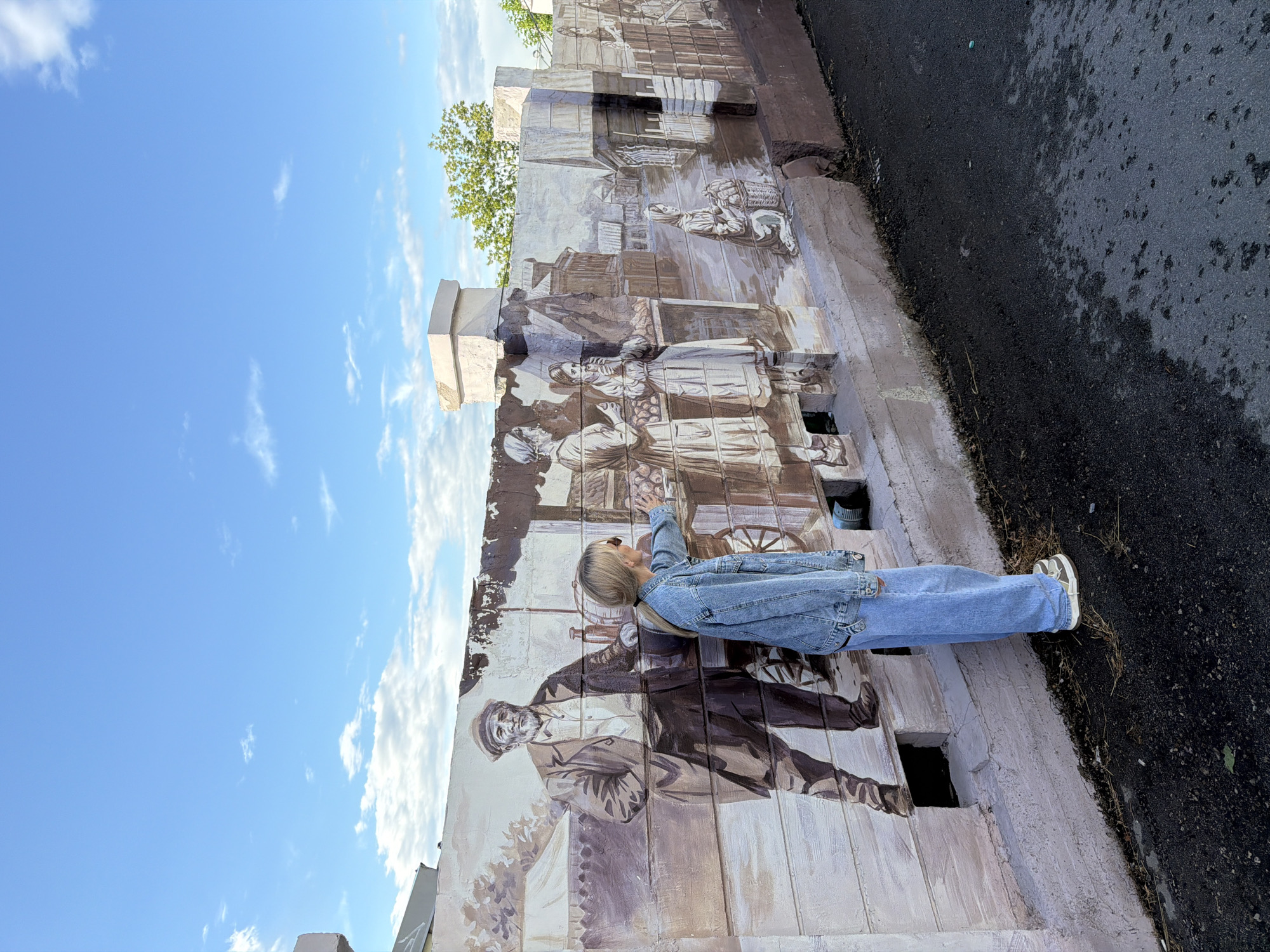 The image depicts a young person, likely female, with blonde hair, wearing a denim jacket and blue jeans, standing and reaching towards a mural on a wall. The mural is rendered in a sepia tone and appears to depict historical scenes with figures in traditional clothing, possibly related to market activities or daily life. One prominent figure in the mural is an older man with a beard, dressed in a suit and hat. The scene is outdoors, with a bright blue sky and scattered white clouds visible. To the right of the mural, there is a dark asphalt surface. The overall impression is a sunny day, possibly in the late morning or early afternoon, given the bright light and clear sky. The mural itself appears to be part of an architectural feature, possibly a building facade or a decorative wall. The person's pose suggests interaction with the artwork, perhaps touching it or pointing at a detail. The location is identified as Nizhny Novgorod, Russia, which adds a cultural and historical context to the mural. There is no visible text in the image.
