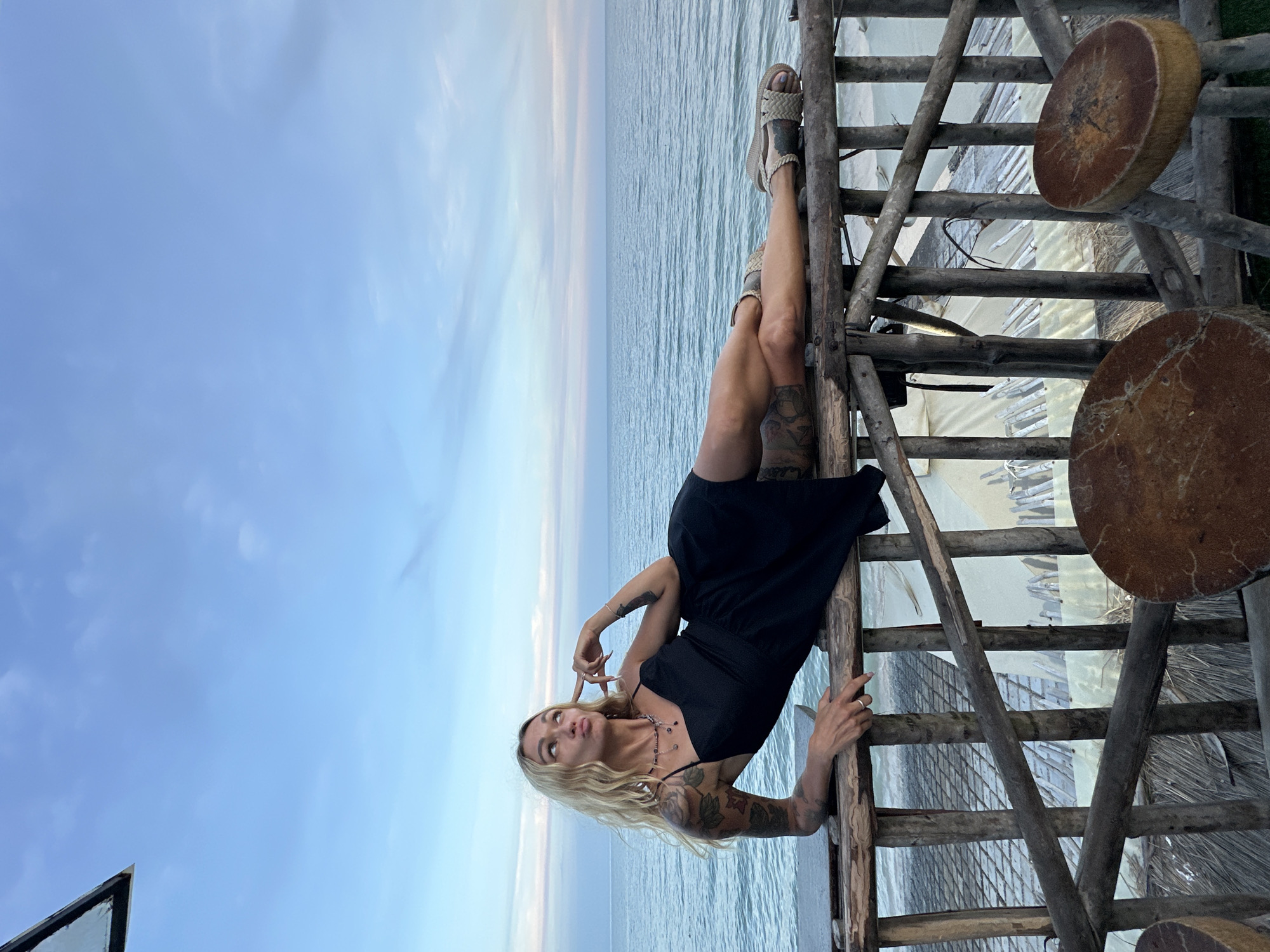 A blonde woman with numerous tattoos is lying on a rustic wooden railing overlooking the ocean. She is wearing a black slip dress and sandals. The sky is a soft blue with wispy clouds, and the ocean is calm. The railing is part of a wooden structure, possibly a balcony or deck, with more wooden posts and some thatched roofing visible below. Two round, rustic wooden stools are positioned in the foreground. The scene appears to be at a beachside establishment in Phu Thuy Ward, Vietnam, likely during the late afternoon or early evening given the lighting.