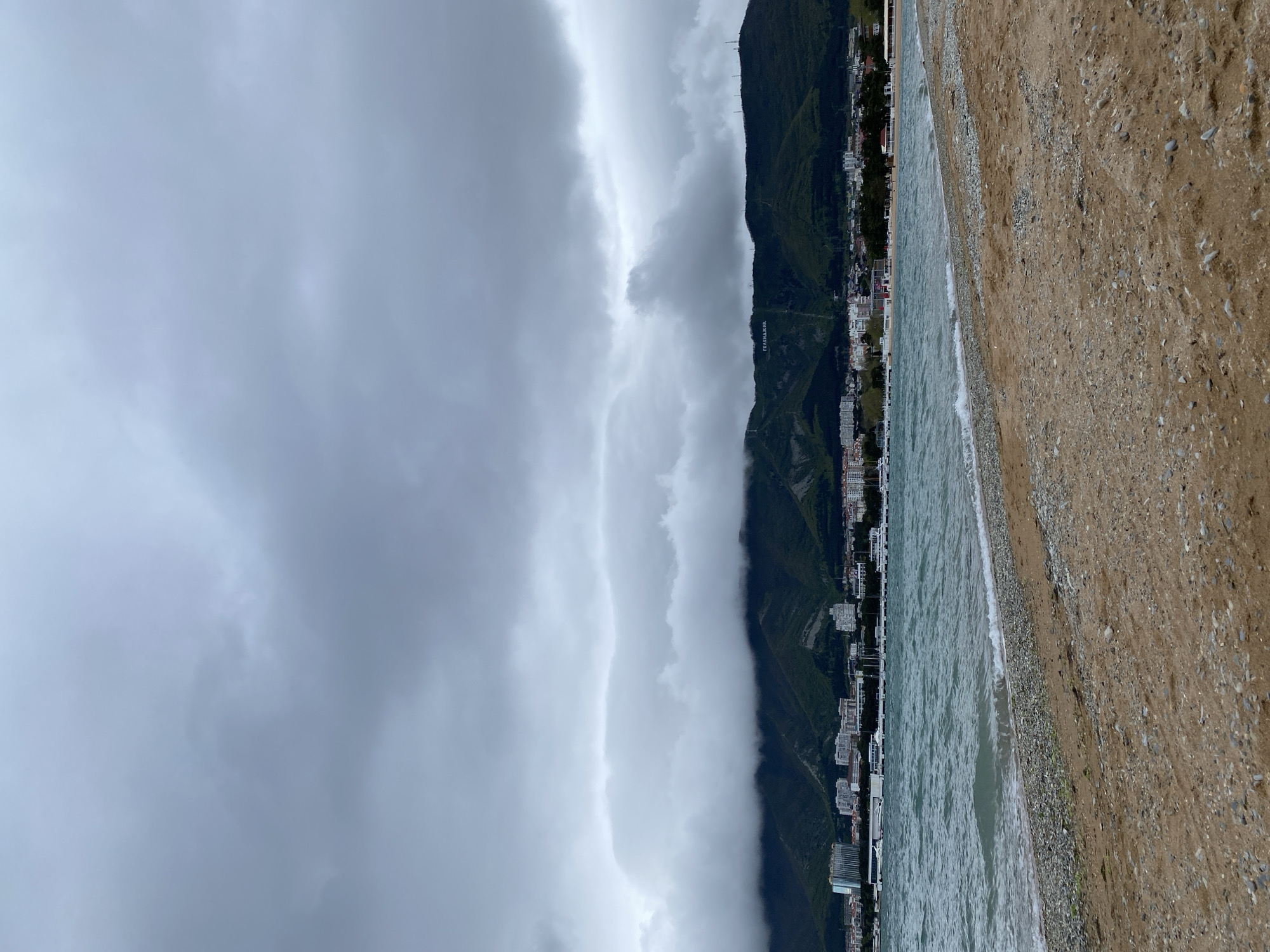 The image captures a view of Gelendzhik, Russia, showcasing a coastline under overcast skies. The foreground presents a beach composed of sand and small stones. Adjacent to the beach is the sea, its surface rippling with small waves. Along the coastline, a line of buildings extends into the distance, nestled between the sea and the base of a mountain range. The mountains rise steeply, their slopes covered in green vegetation. A thick layer of clouds hangs low over the mountains, partially obscuring their peaks and casting a diffused light over the scene. The overall atmosphere is one of a cloudy, possibly stormy, day at the coast.