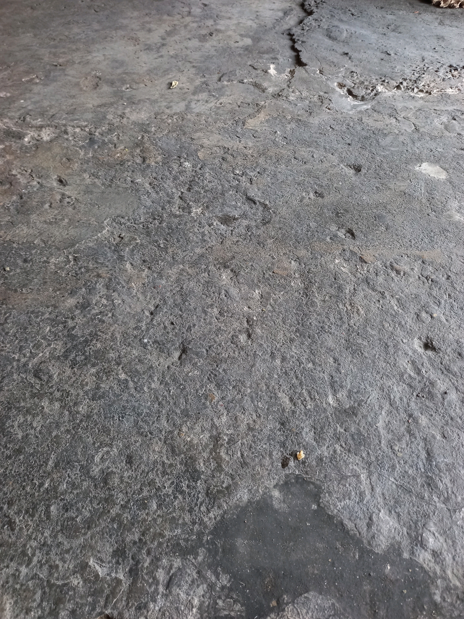 The image shows a close-up, angled view of a rough, dark gray concrete surface, possibly a floor or ground. The concrete appears worn and uneven, with visible cracks and indentations, suggesting age and heavy use. Small specks of debris, likely dirt or gravel, are scattered across the surface. In the upper right portion of the image, there is a more broken and fragmented area of concrete, with lighter colored material exposed. The lighting suggests an indoor or shaded outdoor environment, casting subtle shadows. The scene is devoid of any people or specific objects, focusing solely on the texture and condition of the concrete. Based on the request's context, this scene is located in Samarinda, Indonesia.