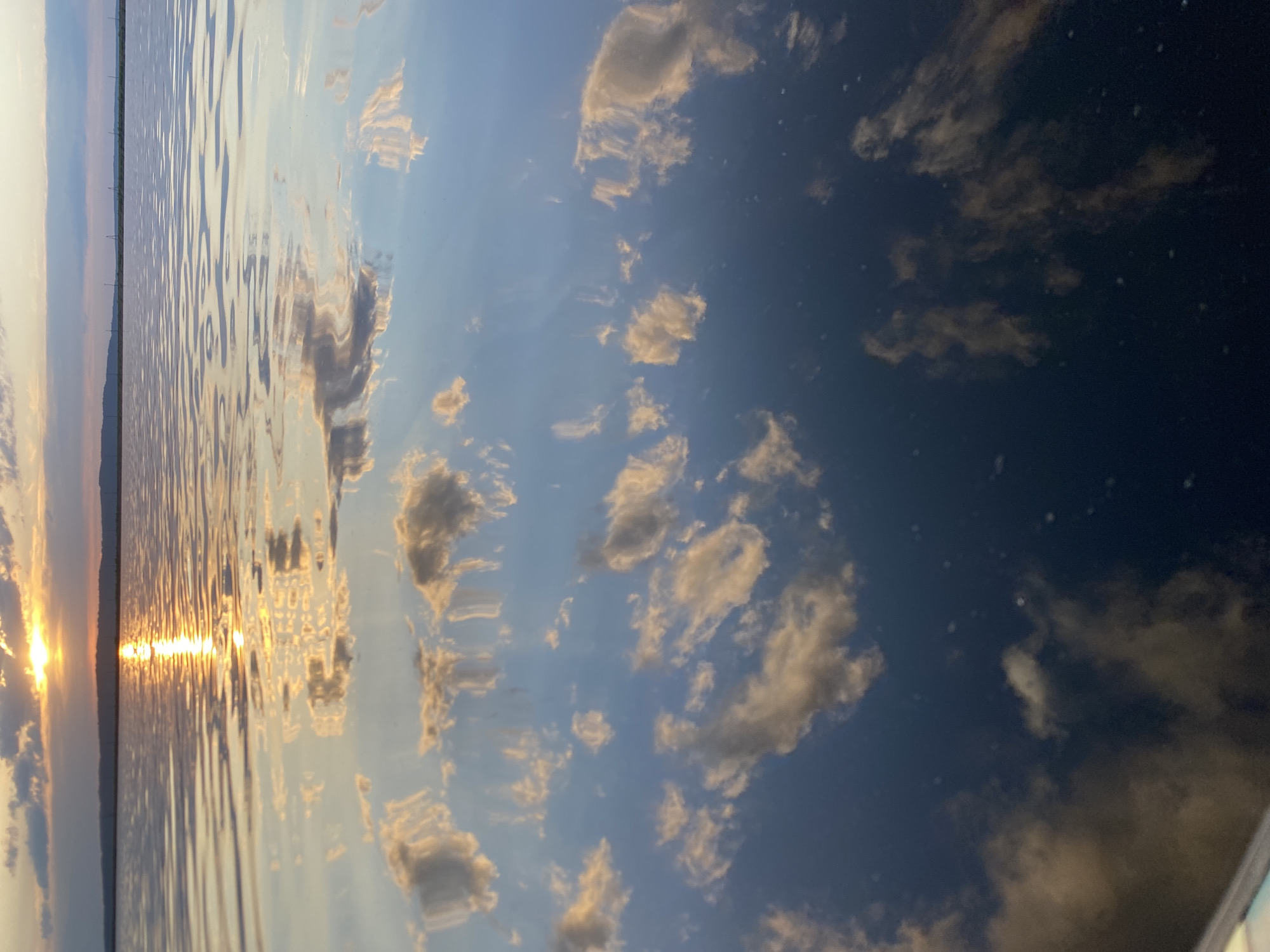 The image depicts a sunset reflected on a body of water, likely a lake or river, near the city of Chaikovsky, Russia. The sky contains scattered clouds, some illuminated by the setting sun. The water's surface is rippled, distorting the reflected light and cloud formations. A dark shoreline is visible on the left, possibly indicating the edge of the body of water or a distant landmass.