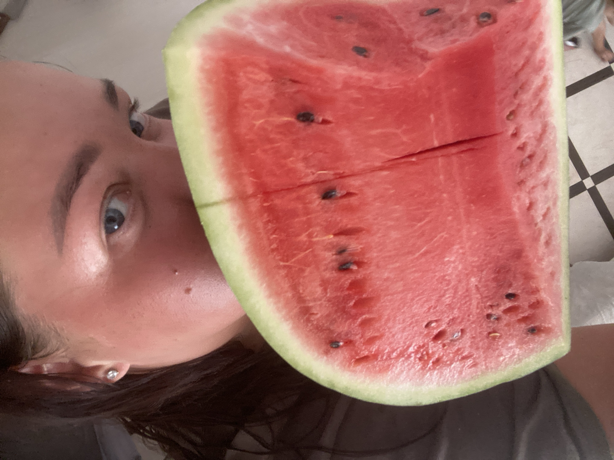 A young woman is taking a selfie while holding a slice of watermelon in front of her face. Her blue eyes are visible over the top of the watermelon. The watermelon is a vibrant red with green rind and black seeds. The background is out of focus, but appears to be an indoor setting with a tiled floor. The image does not provide any information about the location being Sochi, Russia.
