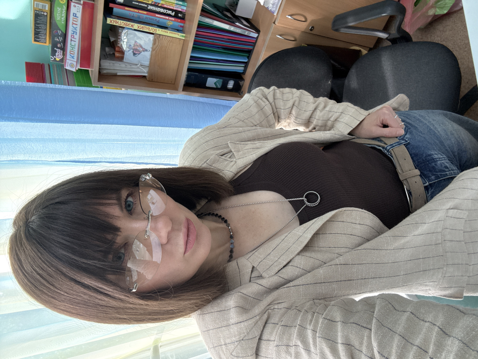 The image depicts a woman in an office-like setting, possibly a classroom or a study room, in Chaikovsky, Russia. The woman, with brown hair and blue eyes, is wearing rimless glasses, a brown tank top, a pinstriped blazer, and blue jeans with a beige belt. She is wearing a necklace with a circular pendant. In the background, shelves are visible, filled with books, folders, and what appear to be educational materials, some of which have Russian text. A blue curtain is also present in the background. A black office chair is partially visible to the right. The overall impression is a snapshot of a woman in her workspace.