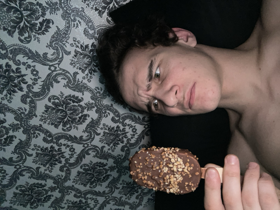 An indoor scene depicts a young adult male within a residential environment located in Krasnoyarsk, Russia. The subject, exhibiting dark, curly hair and light skin, is seen from the upper chest upwards, apparently shirtless. His facial expression displays a furrowed brow, slightly raised eyebrows, and a subtle pout, conveying a look of perplexity or contemplation. In his left hand, he holds a chocolate-coated ice cream bar, generously covered with light-colored chopped nuts, by its wooden stick. His gaze is directed towards the ice cream. The background consists of a wall covered in wallpaper featuring a repeating ornate floral damask pattern in dark grey against a lighter, bluish-grey base. A dark, indeterminate surface is partially visible behind the subject's right side. The scene is lit by an artificial light source, illuminating the subject primarily from the front-right.