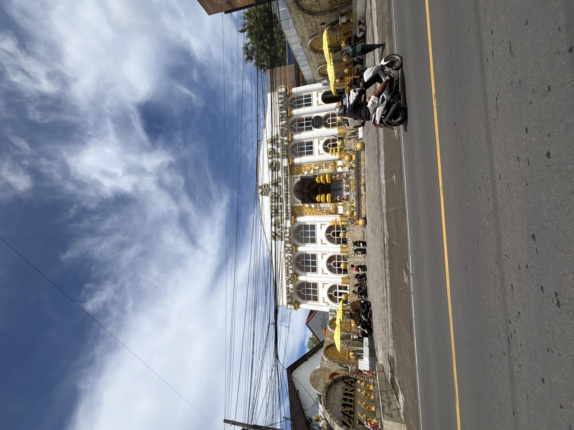The image displays a vibrant street scene in Kuta Selatan, Indonesia, under a bright, partially cloudy sky. The photograph is oriented vertically, showing a road, buildings, vehicles, and people.
The most prominent subject is a large, ornate building with a white facade and extensive golden accents, featuring multiple arched windows and decorative elements. Signs on this building read "THERAPY" and "BEST TATTOO IN BALI" along with "OPEN ALL DAY." Below this main structure, a smaller building with "ZAHRA" inscribed on its stone wall is partially visible. Further back, another small brown sign with "TOKO" is barely discernible.
A paved road occupies the right side of the frame, marked with a clear yellow centerline and a white edge line. Several motorcycles are parked along the sidewalk in front of the buildings. One white and black motorcycle is prominent, with a person seated on it, wearing a white shirt and dark shorts. Another individual stands near a yellow sun umbrella at the building's entrance. The presence of numerous motorcycles suggests a common mode of transport in the area.
The sky is a bright blue, adorned with scattered white, wispy clouds, indicating good weather. Numerous black utility wires crisscross the upper left portion of the image. The scene captures a typical bustling street environment, likely a commercial area, during daylight hours.