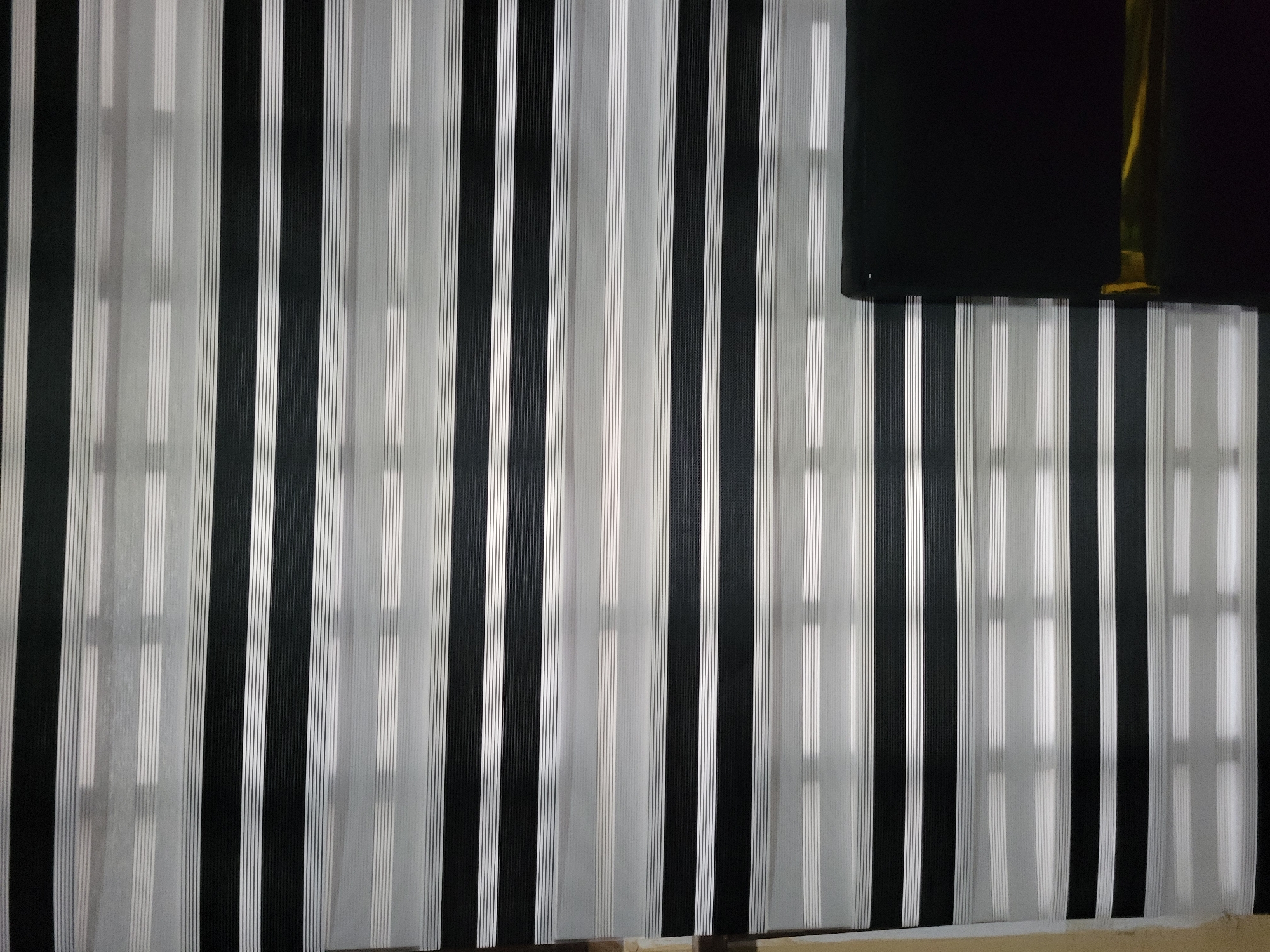An indoor, medium shot captures a section of horizontally striped blinds. The blinds are composed of alternating bands of opaque black and semi-transparent white or grey material, creating a zebra-like pattern of light and shadow. The white/grey bands are further subdivided into thin, parallel lines. The blinds are drawn, partially obscuring the view of what appears to be a window or opening behind them, with faint light filtering through. On the right side of the frame, a portion of a dark, rectangular object, possibly a piece of furniture such as a headboard or a large cabinet, is visible. The object has a reflective golden or brass-colored band near its bottom edge. The scene appears to be in a domestic or commercial interior, with no specific identifying landmarks to pinpoint a location within a city.