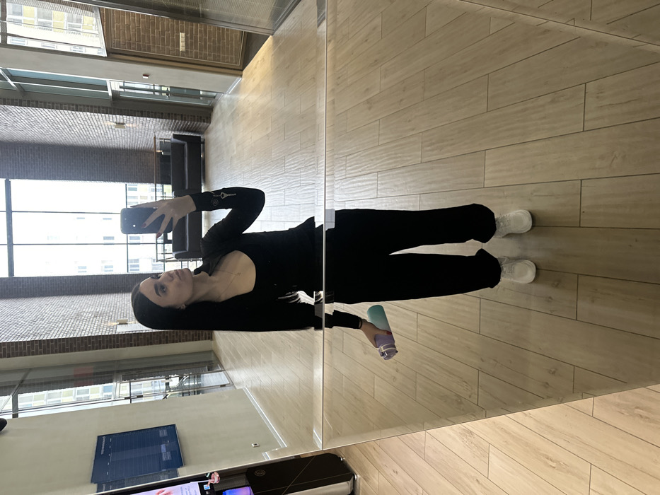 A female individual is captured in a mirror selfie within a modern, high-ceilinged lobby or common area. She has long dark hair and is wearing a black long-sleeved top over a black tank top, black loose-fitting pants, and white athletic shoes. A necklace with a small pendant is visible, as are small key-like charms attached to her right sleeve. She holds a black smartphone in her right hand, pointed towards the mirror, and a purple and light blue/green water bottle in her left hand.
The environment features light-colored wooden or laminate flooring and walls constructed with exposed brick or a brick-patterned facade. Large, floor-to-ceiling windows on the far and right-side walls provide ample natural light and reveal modern multi-story buildings outside, indicative of an urban setting. In the background, two dark-colored sofas are positioned against the far wall. To the left, a tall, vertically oriented digital display screen with blue and white graphics (likely a schedule) stands next to a dark speaker unit with a silver base. The architectural style and internal features are consistent with modern residential, business, or fitness facilities found in Saint Petersburg, Russia, though a precise location within the city cannot be discerned from the image.