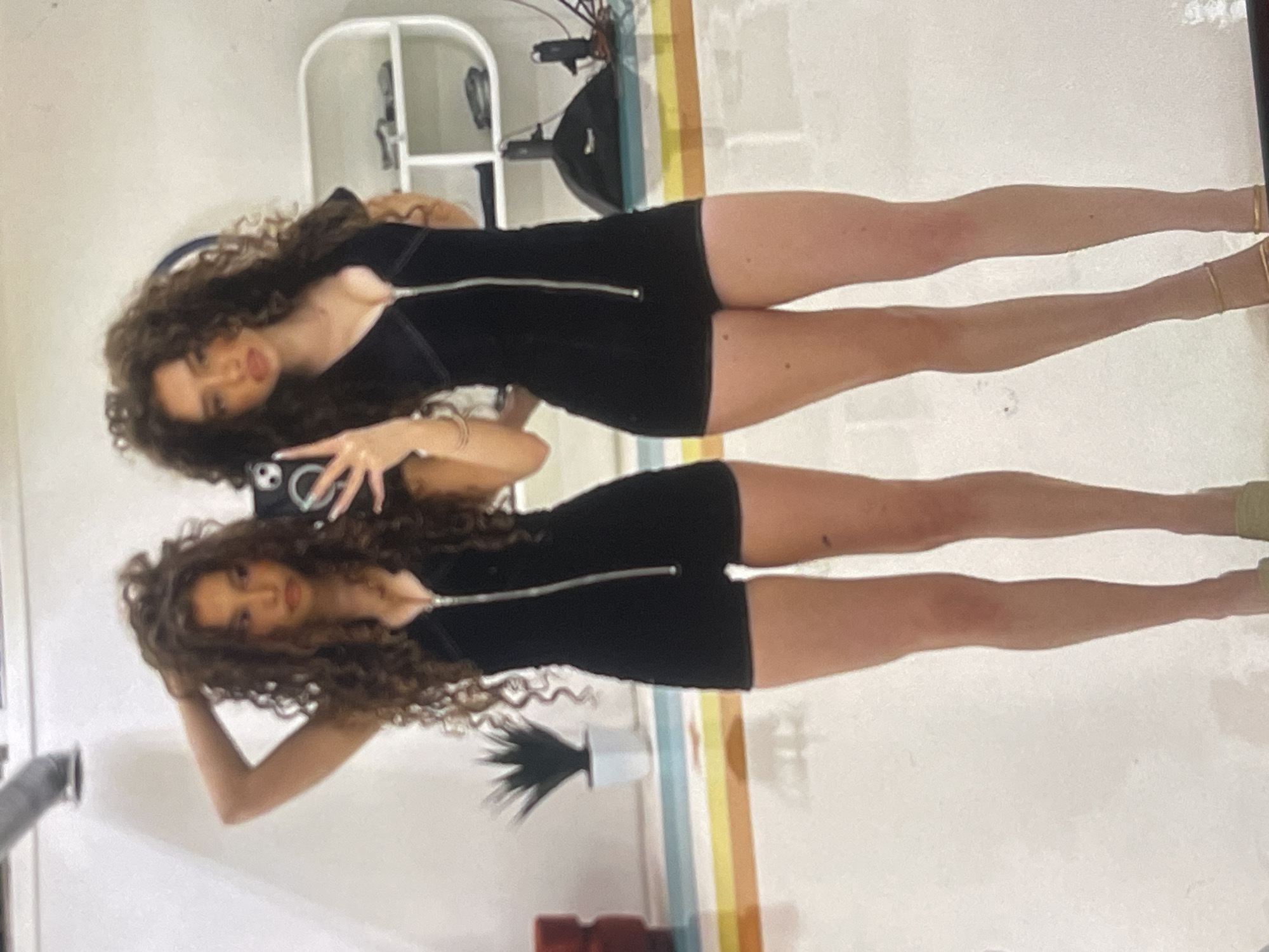 A young woman with long, dark, curly hair is seen twice in a mirror selfie, reflecting herself and her surroundings. She is wearing a fitted black short dress or romper featuring a prominent silver zipper down the front. On her feet, she has gold-colored ankle-strap heels. In the primary reflection, she holds a smartphone (likely for taking the photo) in her raised left hand, while in the secondary reflection, her right arm is bent with her hand near her head.
The background suggests an indoor setting with light-colored walls or flooring. Visible objects include a white shelf or piece of furniture, a dark object that might be gym equipment, and a small plant in a white pot. There's also a decorative element with horizontal stripes in shades of yellow, orange, and light blue or green. The woman maintains a neutral to slightly posed expression. There is no visible text in the image. The scene depicts a casual, well-lit indoor moment, potentially in Poltava, Ukraine.