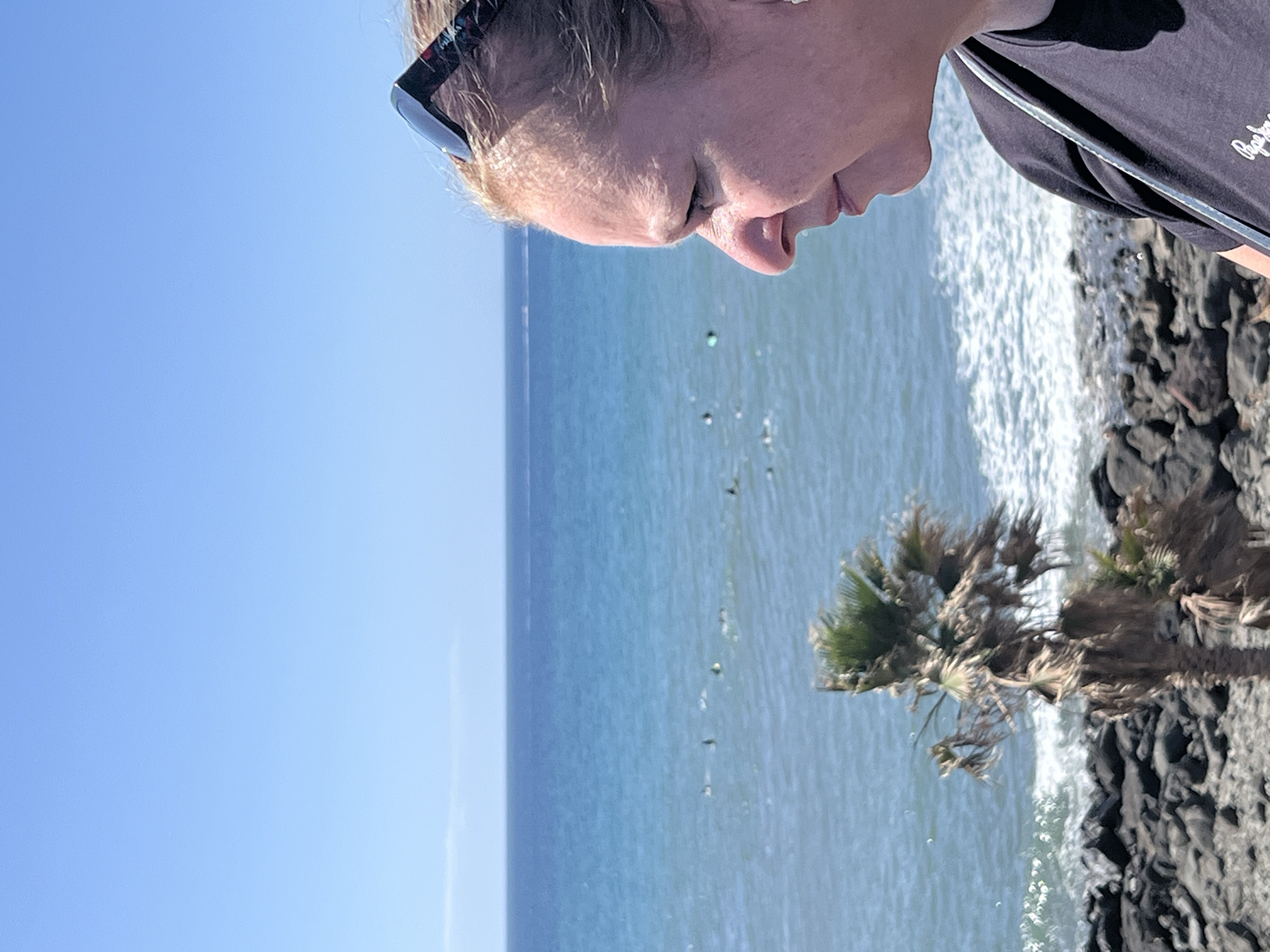 The image depicts a sunny coastal scene in Maspalomas, Spain. A person wearing sunglasses and a dark t-shirt is partially visible in the upper right corner. The camera angle is such that the person is looking down at the ocean.
The ocean stretches across the middle of the frame, appearing light blue and rippled with small waves. A line of the horizon separates the sea from the clear, cloudless blue sky above.
In the lower right, there are dark volcanic rocks along the coastline. A small, windswept palm tree grows near the edge of the rocks. White water from the waves breaks against the rocky shore.
The scene gives the impression of a tranquil day by the sea.
