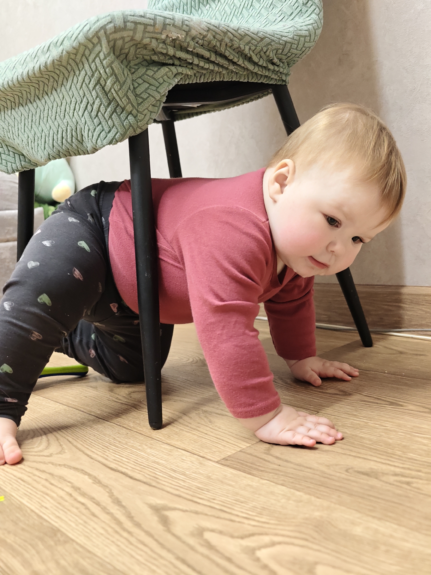 A baby is crawling under a chair. The baby is wearing a pink long-sleeved shirt and dark leggings with small, light-colored shapes printed on them. The chair is covered with a light green, textured fabric. The floor is a light-colored wood. The location is Chaikovsky, Russia.