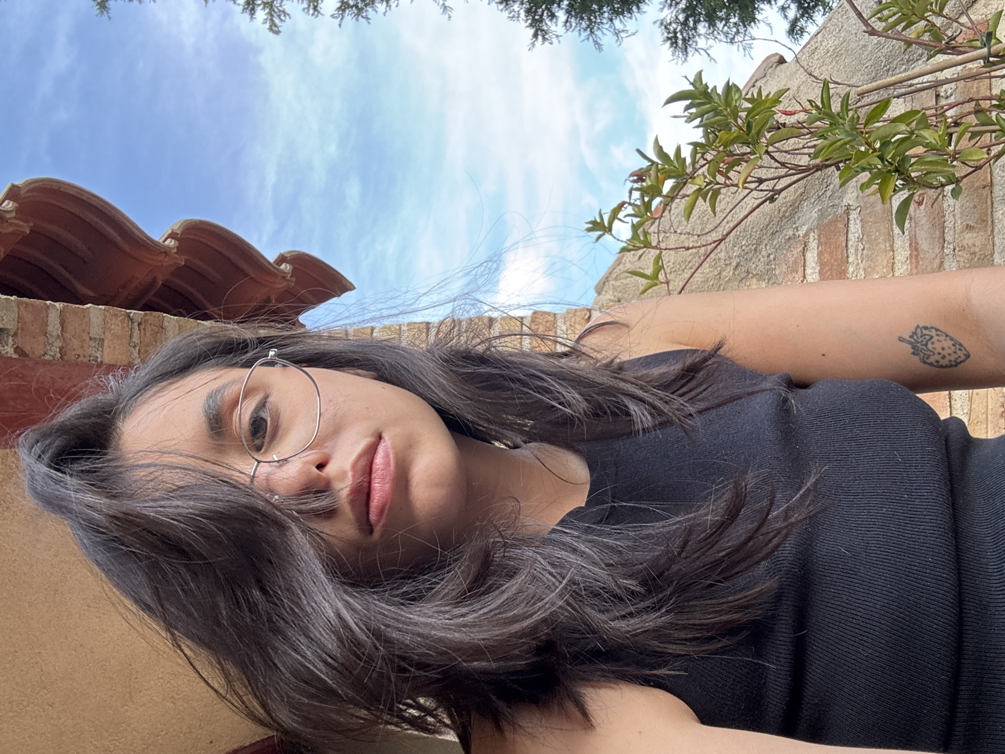 A young woman with long, dark hair and glasses is captured in a close-up, selfie-style photograph. She is wearing a black, ribbed tank top. Her hair partially covers her face, and she is looking directly at the camera with a neutral expression. On her left forearm, a small tattoo of a strawberry is visible. The background consists of a brick wall and a clear blue sky with some clouds. A reddish-brown, tiled overhang is visible in the upper right portion of the image, along with some green foliage. The scene appears to be outdoors, likely on a balcony or patio, given the architectural elements and the presence of plants. There are no other individuals or significant objects in the frame. The photograph was taken in Las Rozas de Madrid, Spain.
