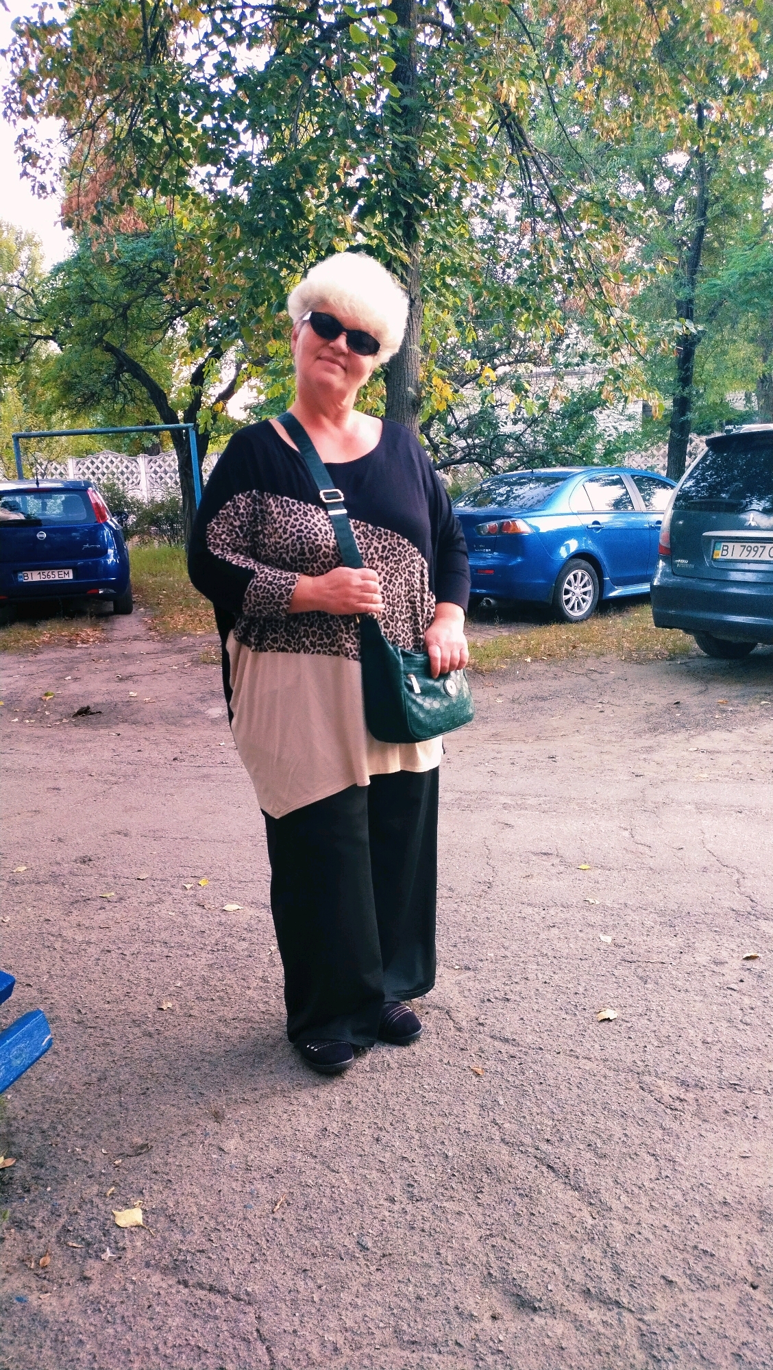 A woman stands outdoors in a parking lot in Horishni Plavni, Ukraine. She has fair skin and blonde hair, and is wearing dark sunglasses and a black top with a leopard print pattern across the chest, a tan undershirt, and black pants. She is carrying a small, dark green purse with a shoulder strap. Behind her, several cars are parked, including a blue sedan and a dark colored SUV. Trees with green and brown leaves are visible in the background, suggesting it is autumn. The ground is paved with asphalt and scattered with a few fallen leaves.