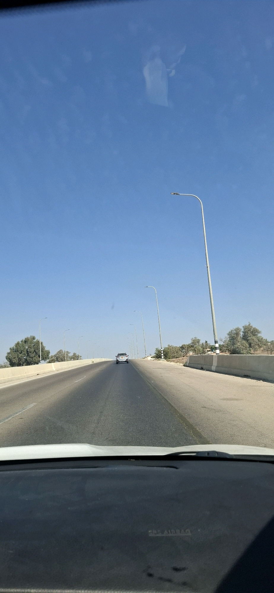 The image depicts a clear, sunny day with a bright blue sky and a few wispy clouds. The perspective is from inside a vehicle, looking out through the windshield and dashboard. A paved road stretches into the distance, with a single car visible ahead. Streetlights line the right side of the road, and sparse vegetation, including trees and shrubs, dots the landscape on both sides. The scene appears to be on a highway or a significant road outside of a city center, given the open space and the presence of streetlights and a well-maintained road surface. The environment suggests a dry, possibly arid climate. The text "SRS AIRBAG" is visible on the dashboard, indicating it is a modern vehicle. The landscape suggests a location in the Neot Hovav area of Israel, known for its industrial zones and desert surroundings.