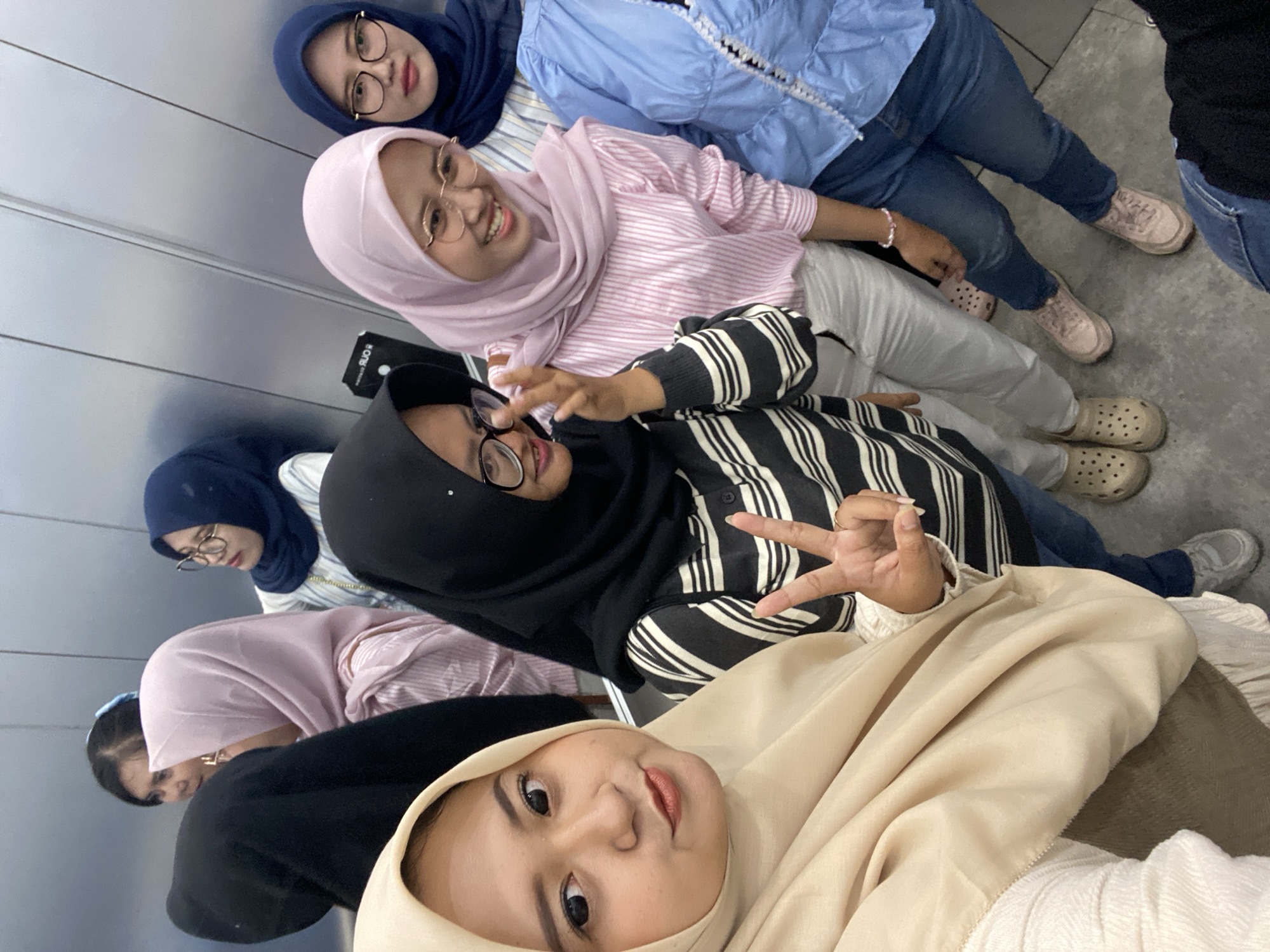 A selfie photograph shows six women in Sidoarjo, Indonesia, wearing hijabs in varying colors. From the front, one woman is wearing a beige hijab and white sweater. Behind her, a woman with glasses is wearing a black hijab, striped shirt, and making a peace sign with her right hand. A third woman wears a pink hijab, a pink and white shirt. The remaining three women in the back wear navy blue, navy blue, and light blue hijabs, respectively. The women are wearing glasses and varying colored shoes. They are in an elevator with brushed metal walls and floor.
