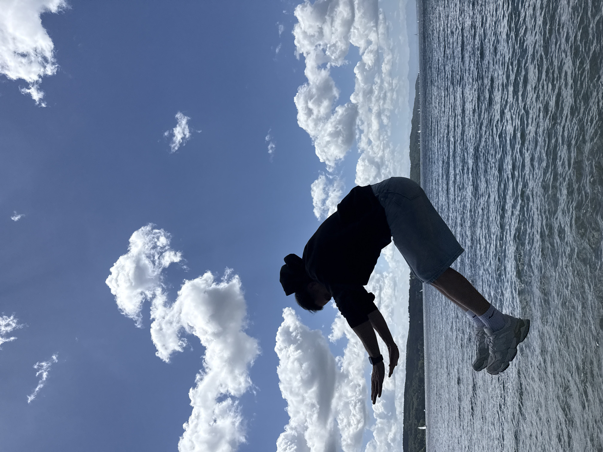 A person in silhouette is captured mid-air, arms outstretched, in front of a body of water and a blue sky with white clouds. The person is wearing a dark hooded sweatshirt and denim shorts. Their legs are bent, and they are wearing light-colored sneakers and white socks. The water appears choppy, with ripples and small waves. In the distance, a treeline with hills and mountains can be seen, with several sailboats on the water. The scene is likely Starnberger See, Germany, given the presence of sailboats and the overall landscape.