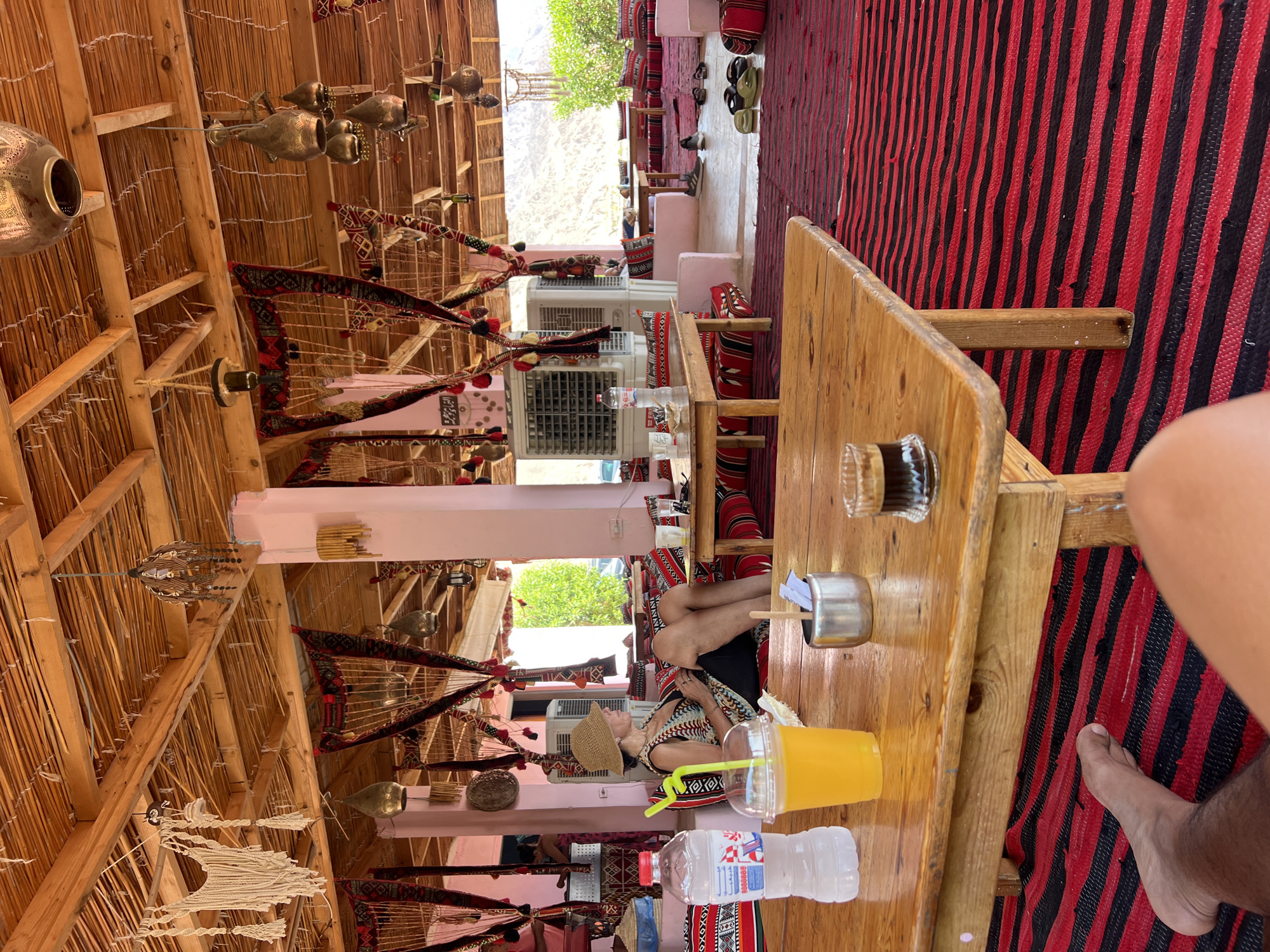 An outdoor covered seating area with a thatched roof and pink pillars is depicted. The floor is covered with a red and black striped rug. In the foreground, a wooden table holds a bottle of water, a cup of orange juice with a straw, a metal sugar dispenser with packets, and a small glass of dark liquid, possibly coffee. A person's legs and feet are visible in the bottom left and center of the frame. In the mid-ground, a woman wearing a straw hat and patterned vest is seated on a cushion, looking to her right. Behind her, more seating areas with cushions are visible, along with large air conditioning units mounted on the wall. Decorative tapestries with red, black, and gold patterns hang from the ceiling and pillars. In the background, there is greenery and a mountainous landscape under a bright sky. The overall atmosphere suggests a relaxed, possibly tourist-oriented, establishment in a desert region.