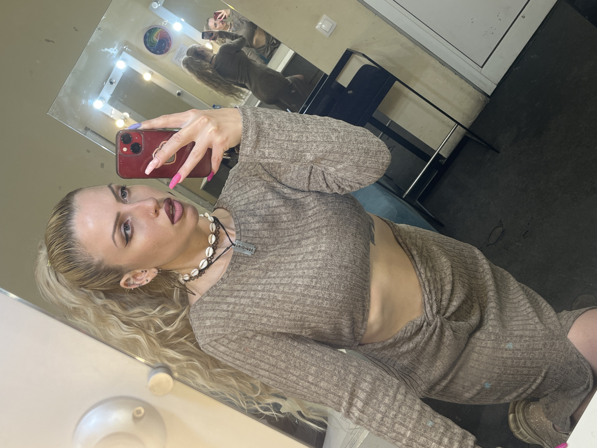 A woman is taking a selfie in a room that appears to be a backstage or dressing room, possibly in a theater or similar performance space. The location may be Plovdiv, Bulgaria. The woman has blonde hair and is wearing a brown, long-sleeved dress with cutouts at the side. She is holding a red mobile phone, capturing her reflection in a mirror. She has multiple piercings in her ear and is wearing a necklace with what appear to be shells and a small metal charm. In the mirror's reflection, another person with blonde hair is visible, possibly taking a picture as well. The room features a mirror with lights around it, a chair, and a portion of a door or wall, suggesting an interior space.