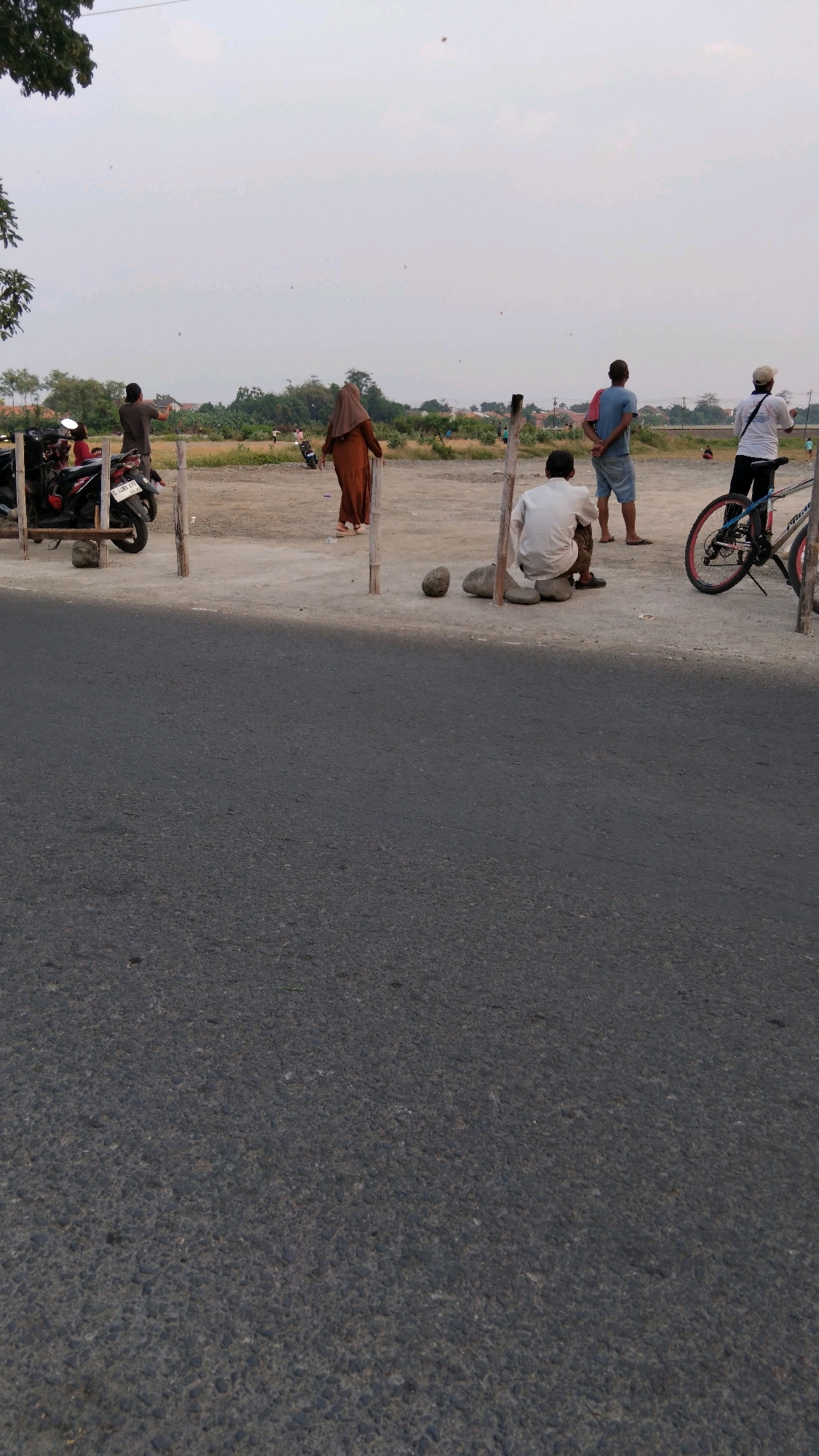 In Tegal, Indonesia, a group of individuals are gathered near a dirt road and a paved street. The sky is overcast and light. In the foreground, the textured asphalt of a street dominates the lower portion of the image. The middle ground features a dirt lot with several wooden posts, suggesting a makeshift barrier or structure. Several motorcycles are parked to the left.
To the right of the motorcycles, a woman in a brown hijab and dress walks away from the camera, looking towards the sky. Further back, a person in a dark t-shirt stands facing away, also looking skyward. Another individual in a blue shirt and shorts stands with their back to the camera. In the center of the dirt lot, a person in a white shirt sits on rocks, facing away from the viewer. A bicycle with red accents is parked to the right. In the distance, a treeline and some buildings are visible, indicating a rural or suburban setting. Several small, distant objects are visible in the sky, possibly kites. The overall scene appears to be a casual gathering, possibly for recreational purposes like kite flying.