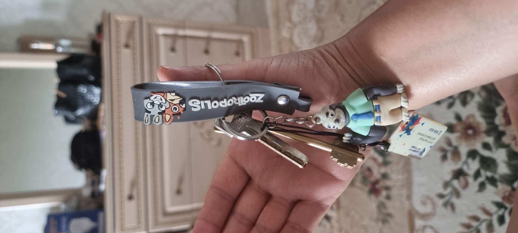 In this close-up image, a hand is holding a set of keys, including a large, antique-looking brass key and several smaller keys. Attached to the keys is a gray keychain with the "ZOOTROPOLIS" logo and characters from the movie. Below the keychain, a small figurine of a sloth character from Zootropolis, wearing a light blue shirt and a darker blue tie, hangs from a separate chain. A small white tag with Cyrillic text, possibly an identification or delivery tag, is also attached. The background is blurred, showing a dresser or cabinet with drawers, suggesting an indoor setting. A faint floral pattern is visible on the surface below the hand. The image is a shallow depth of field, focusing on the keys and the hand.