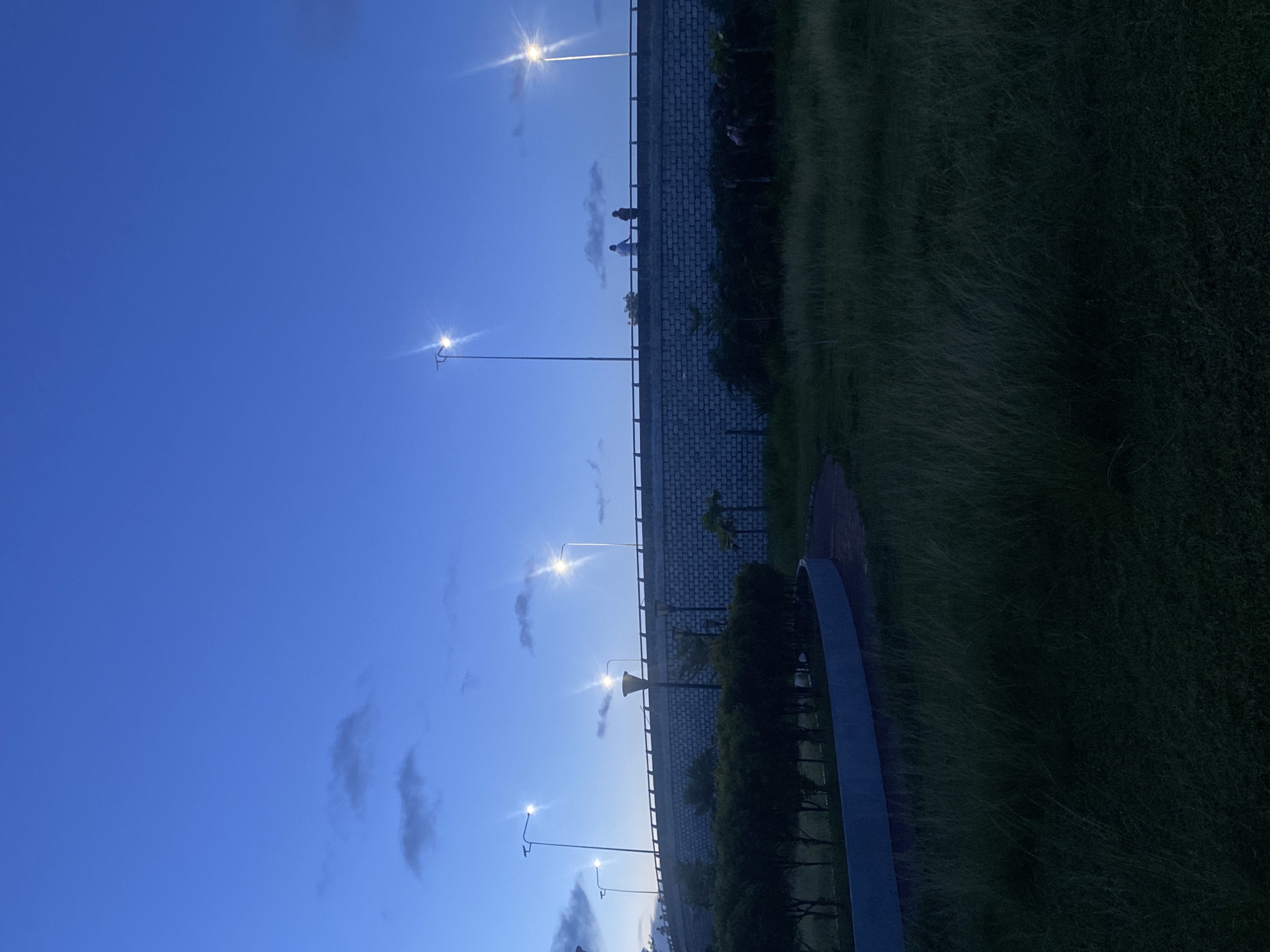 The image captures a twilight scene in Davao City, Philippines, with a deep blue sky overhead, sparsely dotted with faint clouds. In the foreground, a grassy field stretches towards a curved pathway and a manicured row of dark green bushes. Beyond the bushes, a substantial concrete wall, constructed of light gray bricks, forms the base of an elevated structure, possibly a bridge or overpass. Several tall lampposts with bright lights are strategically placed along this structure, illuminating the area. Two figures are visible standing on the elevated walkway, silhouetted against the brightening sky, appearing to observe the surroundings. The overall atmosphere suggests a public park or recreational area.