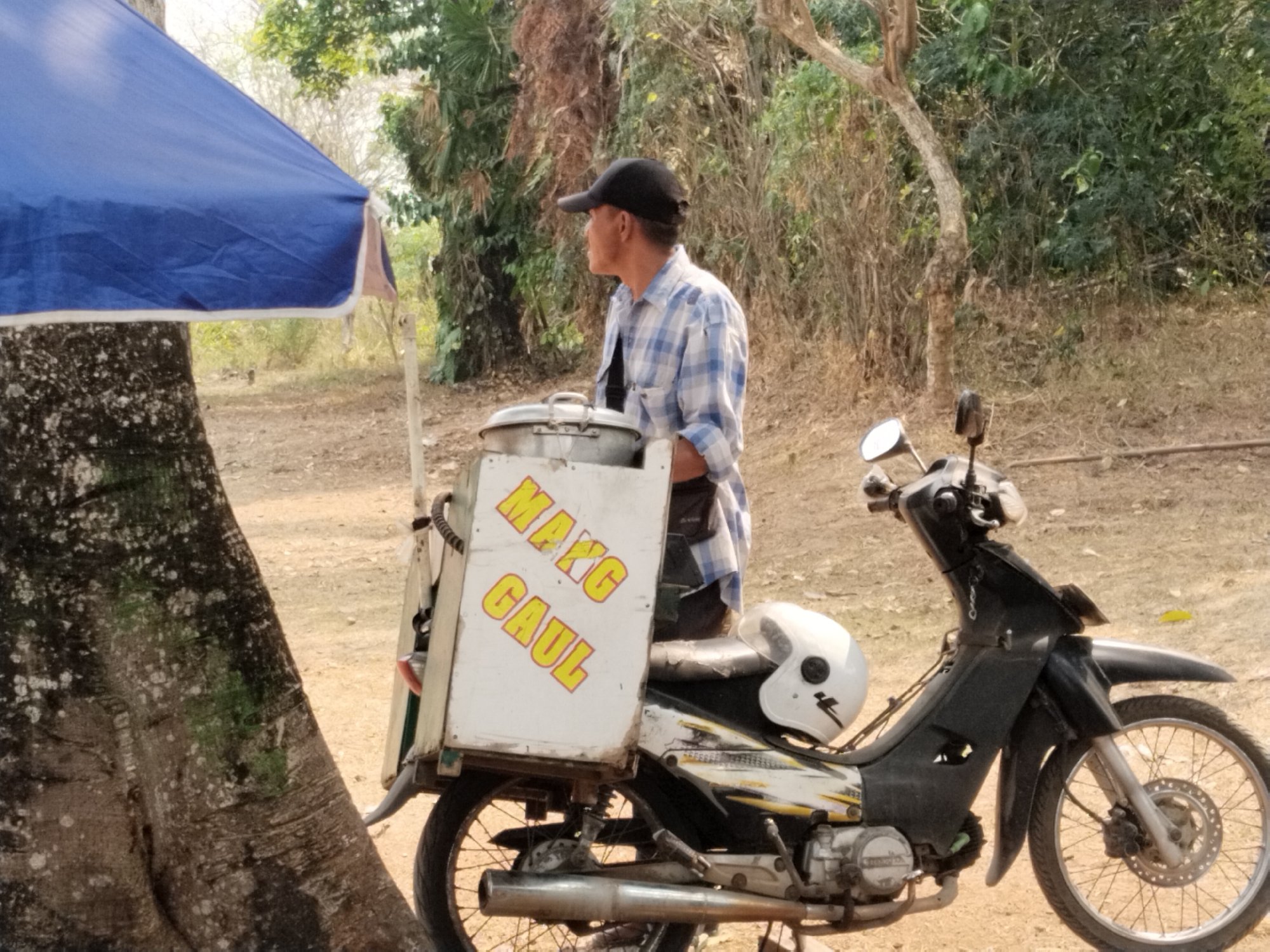 The image depicts a street vendor on a motorcycle in Serang, Indonesia. A man wearing a blue and white checkered shirt and a black baseball cap is standing next to his motorcycle, which is equipped with a food cart. The food cart has the words "MANGO GAUL" printed in yellow on a white background. A white helmet with a black sticker is placed on the seat of the motorcycle. The background shows a dirt road, trees, and foliage, suggesting a rural or semi-urban setting within Serang. The overall scene appears to be a typical street view of a vendor preparing for or concluding business.