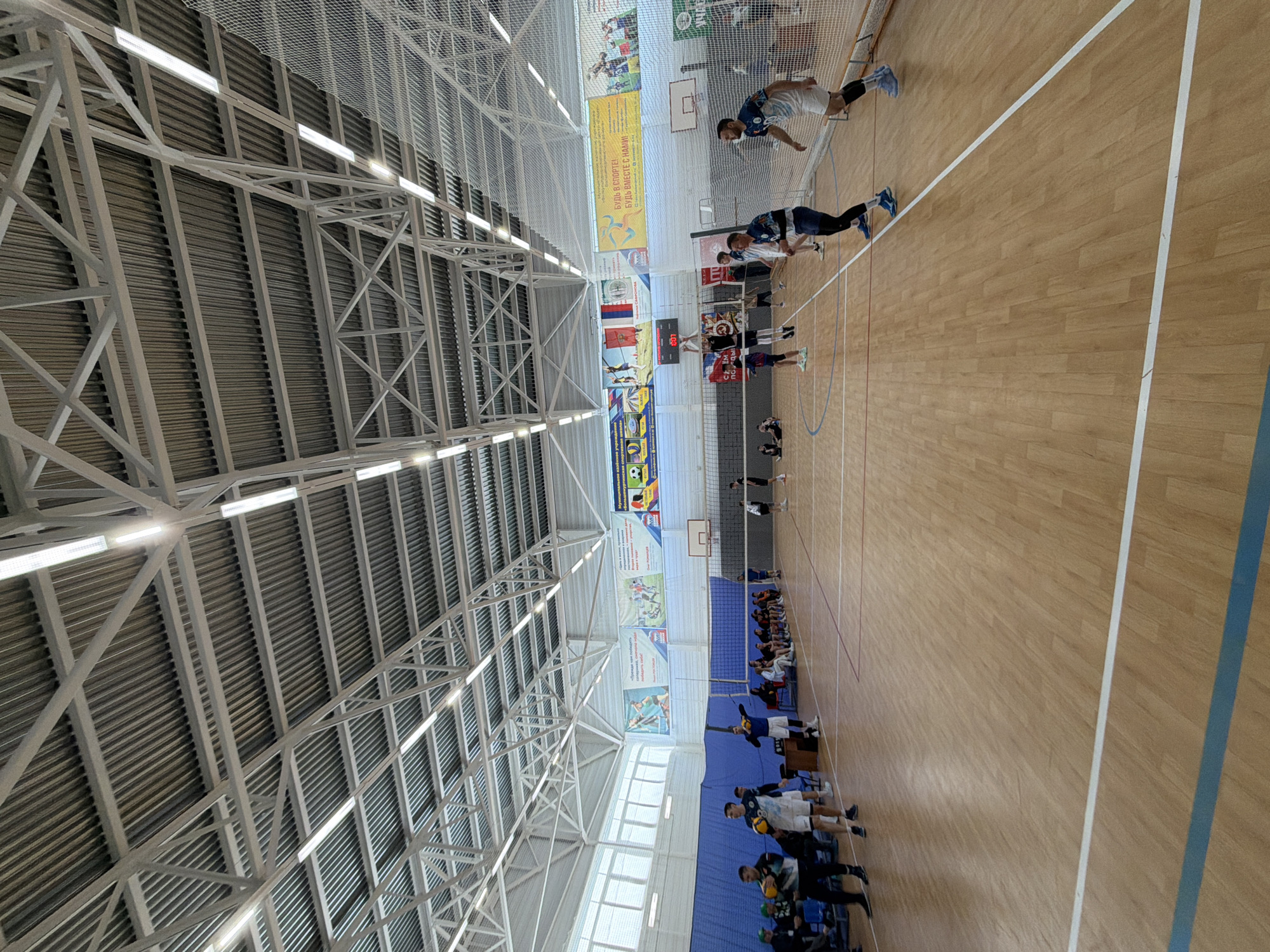 The image shows a volleyball game in progress in a gymnasium. The gymnasium is located in Bolshaya Murta, Russia. The gymnasium has a high, metal-supported ceiling. A volleyball net divides the court. Two teams are playing volleyball. Players in dark blue uniforms are on the near side of the net. Players in light blue uniforms are on the far side of the net. There are people sitting in the bleachers watching the game. Banners adorn the walls of the gymnasium.