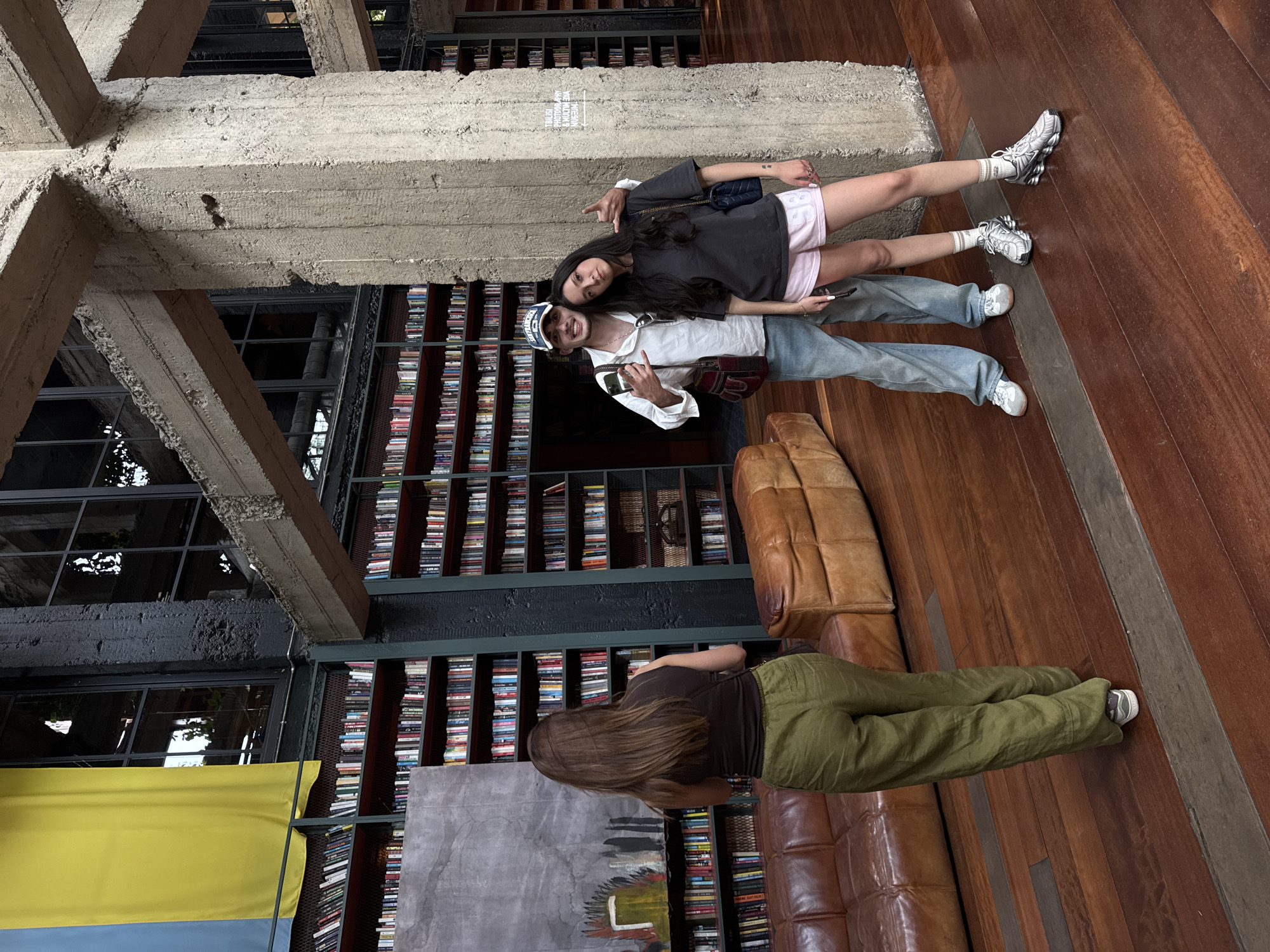 In a large, industrial-style room with exposed concrete beams and pillars, a Ukrainian flag hangs prominently on the left side of the frame. The walls are lined with floor-to-ceiling bookshelves filled with numerous books. In the foreground, a woman with long brown hair, wearing a dark tank top and olive green pants, stands with her back to the camera, looking towards the bookshelves. To her right, a man wearing a white long-sleeved shirt, blue jeans, and a baseball cap, and a woman in a dark t-shirt and pink shorts, pose for a photo. The man has his arm around the woman, and both are smiling. A brown leather sofa is visible in the background between the woman in green and the couple. The floor is made of dark polished wood. The location is the Tbilisi Photography and Multimedia Museum in Tbilisi, Georgia, as indicated by a sign on a concrete pillar in the background.