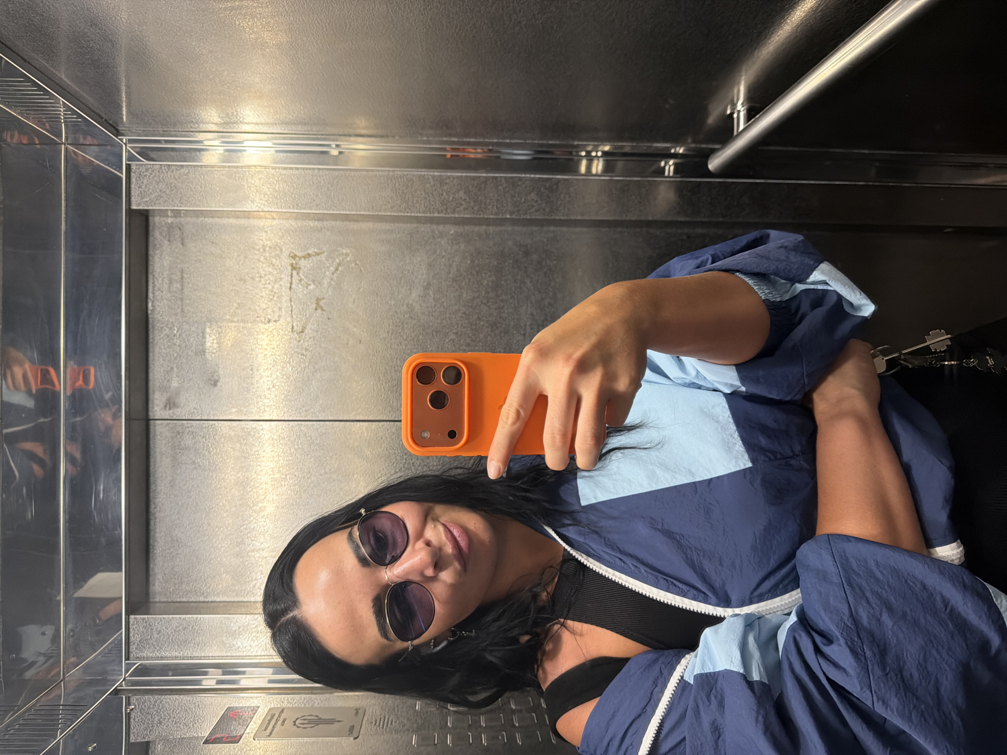 A woman is taking a selfie in an elevator in Saint Petersburg, Russia. She has long, dark hair and is wearing round, purple-tinted sunglasses and a blue and light blue windbreaker jacket over a black top. She is holding an orange smartphone in her right hand, which is angled to capture her face and upper body. The elevator walls appear to be made of brushed metal, with a handrail visible on the right side of the frame. The lighting suggests it is daytime. The woman has a slight smile and appears relaxed. There are some smudges and reflections on the elevator walls. In the lower part of the frame, there are two small cards visible on the wall, likely indicating elevator regulations or information, but the text is too small to be legible.