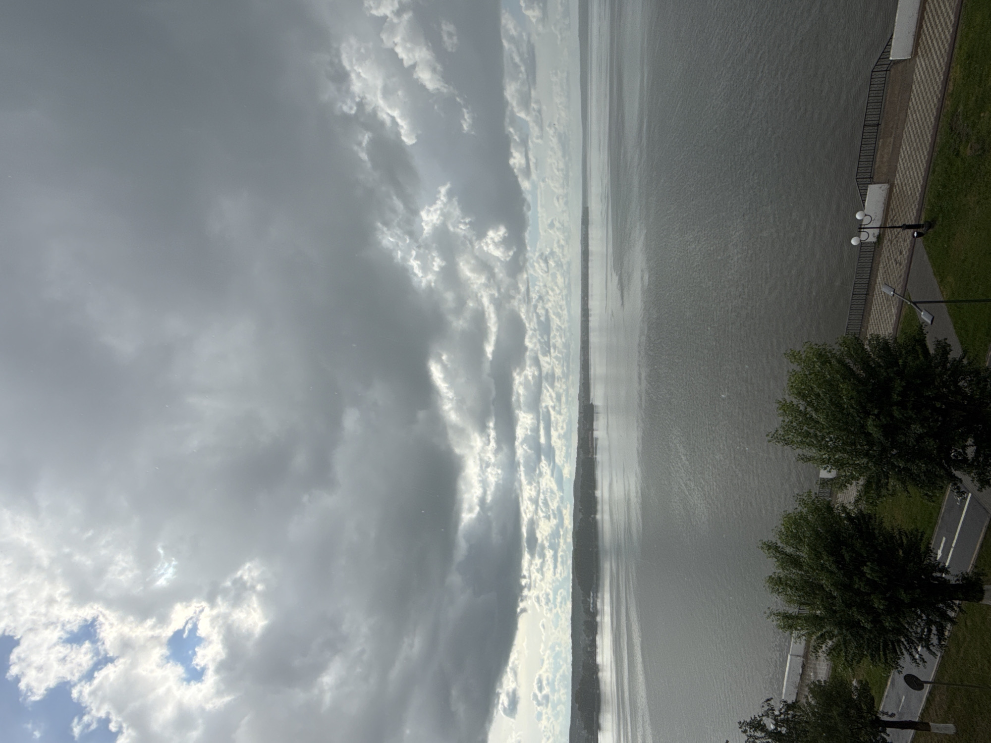The image depicts a waterfront scene in Votkinsk, Russia. The sky is filled with heavy, predominantly gray clouds, but with patches of light blue peeking through. Below the sky, a large body of water fills most of the frame, with subtle ripples visible on its surface. Along the right side of the image, there is a paved walkway and some green grass. On the walkway, there are lampposts and a section of wrought-iron fencing. Some trees line the walkway. A paved road or path runs behind the trees.