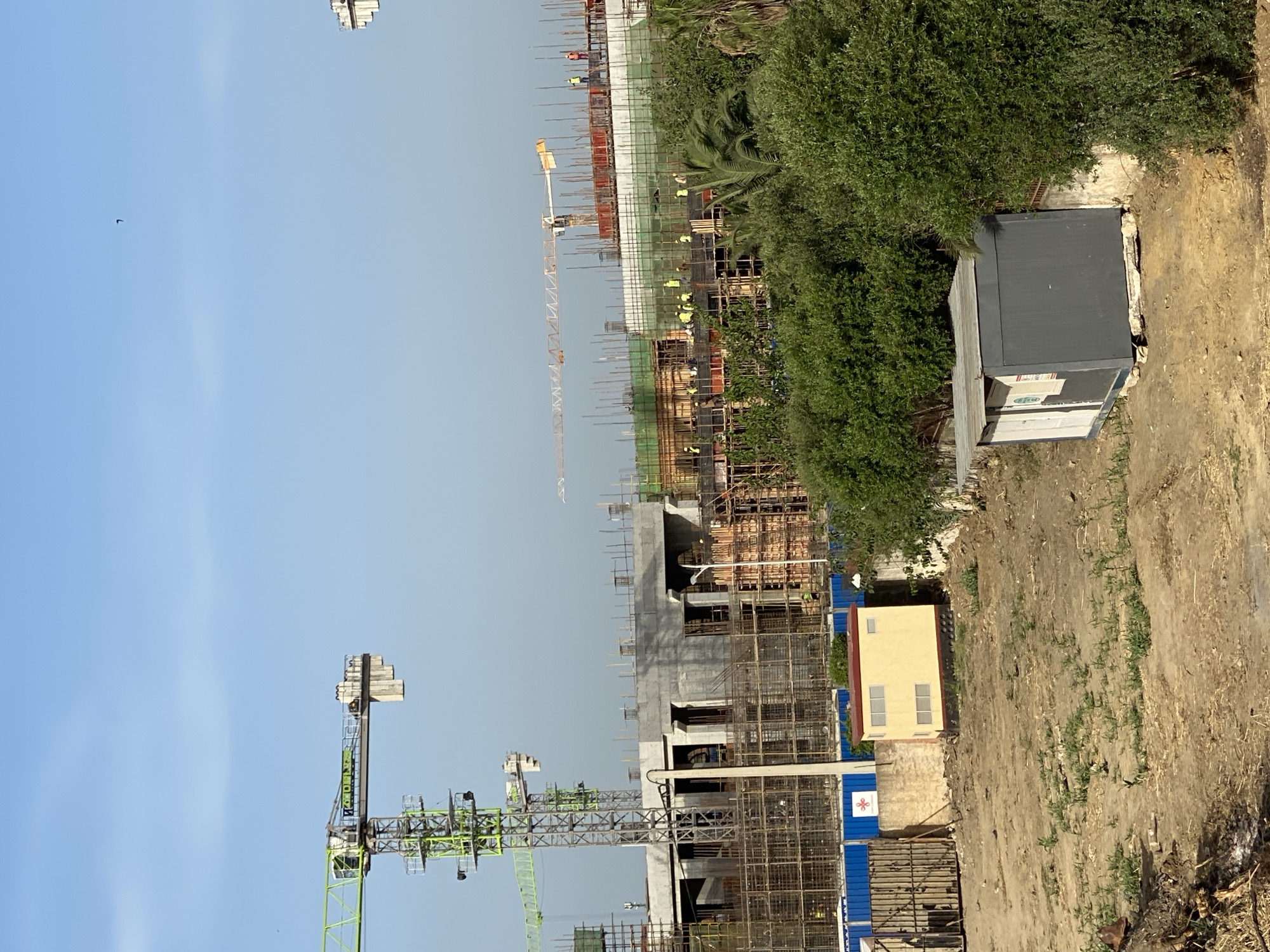This is a full, eye-level, outdoor, daytime shot of a construction site in Baraki, Algeria. The scene is dominated by a large concrete building under construction, with scaffolding and construction equipment surrounding it. In the foreground, there is a dirt area with some sparse vegetation, a small yellow building with red trim, and a dark gray portable cabin. In the background, several construction cranes, including a prominent green one on the left and a yellow one in the center, are visible against a clear blue sky. The construction site appears to be active, with workers visible on the upper levels of the building. The overall impression is one of ongoing development and industrial activity in an urban setting.