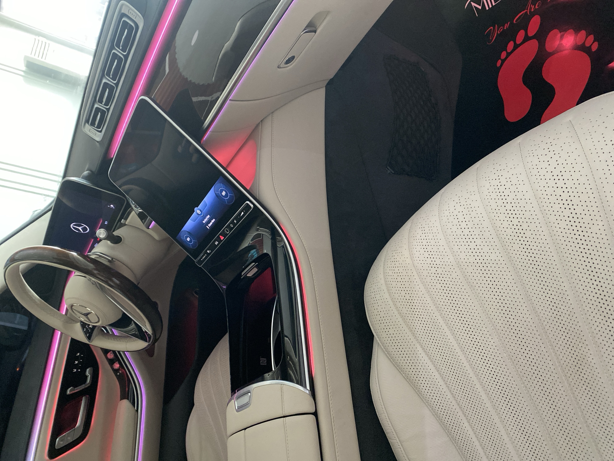 The image shows the interior of a car, likely a Mercedes-Benz, given the steering wheel emblem. Key features include a digital dashboard display, a white leather seat with perforations, ambient lighting (pink/red), and a black mesh pocket behind the seat. There's a red footprint graphic in the background. The location is not discernible based on the image content alone.