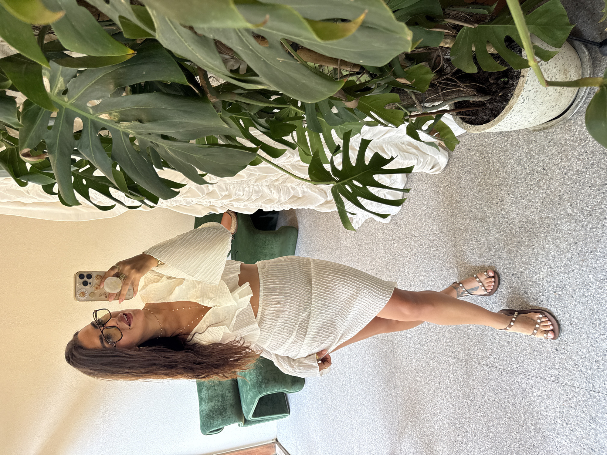 A woman in a white dress and glasses stands on a terrazzo floor in Kuta Selatan, Indonesia, taking a selfie with a phone. She is positioned in front of a green velvet chair and a large potted Monstera plant. The plant sits partially on top of a white draped object, possibly a bed or table. The woman's sandals are visible. The background is a combination of a light-colored wall and the terrazzo floor.
