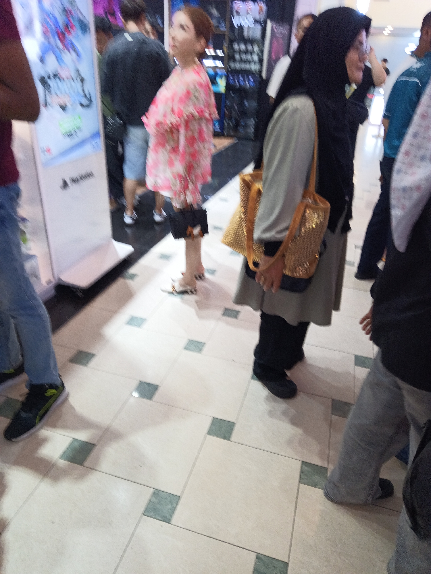The image shows a street-level view of people walking indoors, likely in a commercial area in Kuala Lumpur, Malaysia. A woman in a pink floral top and dark skirt is in the foreground, holding a gold purse. To her right, a woman wearing a black hijab and grey long-sleeved shirt also holds a gold purse. To the left, a person wearing blue jeans and a black and green shoe is partially visible. Another person is seen wearing grey pants and black shoes. The environment appears to be a tiled walkway with some retail displays in the background. The lighting suggests an indoor setting with artificial illumination.