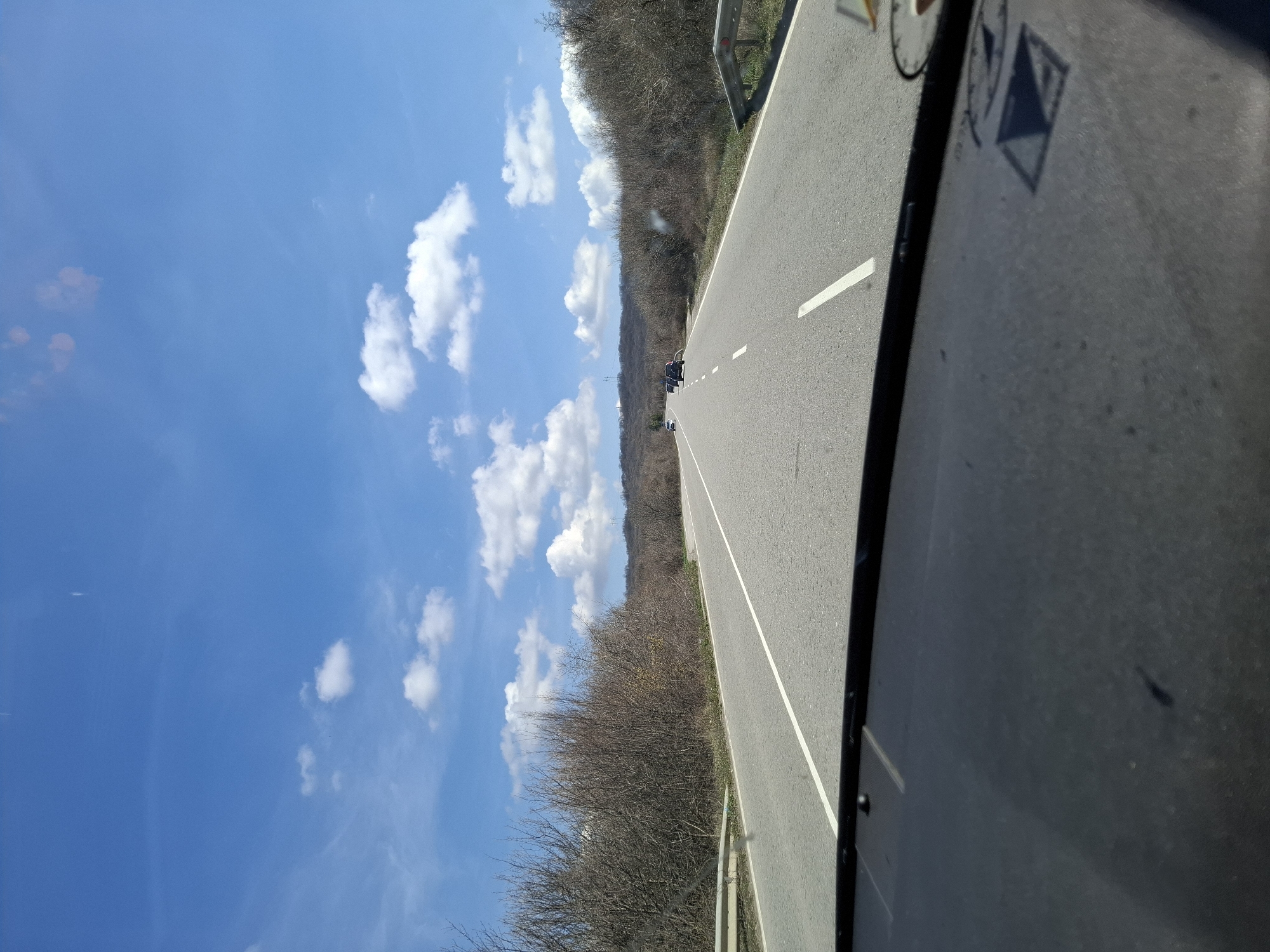The image shows a two-lane road in Bogyovtsi, Bulgaria, flanked by dry, leafless trees on either side. The sky is blue with scattered white clouds. A vehicle is visible ahead on the road, along with a few other vehicles in the distance. The image is taken from inside a vehicle, with part of the vehicle's interior visible on the right side of the frame.