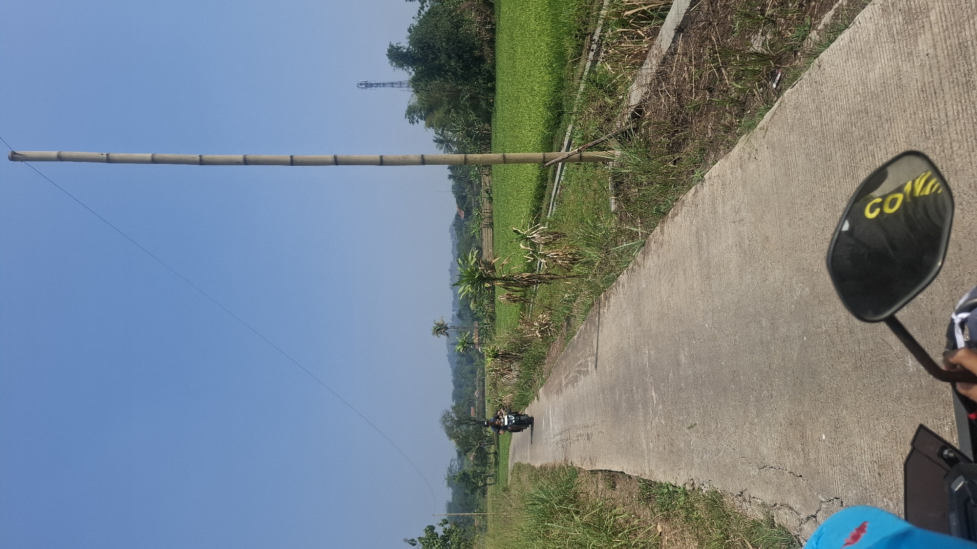 This is a perspective shot from a motorcycle on a concrete road in Cianjur, Indonesia. A person is riding a motorcycle in front, on the same road, heading away from the camera. The road is lined with dry grass and some green vegetation. To the right of the road, a large field of green crops, likely rice, stretches into the distance. In the background, there are trees, a few houses, and a cell tower. A tall, slender bamboo pole stands on the right side of the road, with a wire extending diagonally upwards and to the left across the clear blue sky. A cell phone tower is visible in the far distance behind the bamboo pole. The motorcycle's rearview mirror is in the foreground, reflecting the road and some yellow text. The overall scene depicts a rural landscape under a bright, sunny sky.