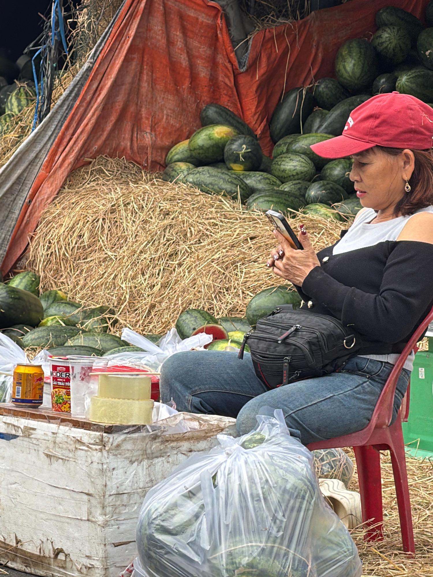 A woman wearing a red baseball cap and a black off-the-shoulder top is sitting on a red plastic chair and looking at her phone. She is wearing blue jeans and has a black fanny pack around her waist. She is at a roadside fruit stand in Nha Trang, Vietnam, selling watermelons. The stand is covered by a large orange and grey tarp, and there is a large pile of hay behind the watermelons. In front of the woman, on a makeshift wooden table, are two cans, a cup, and some clear containers. Several plastic bags filled with watermelons are also visible. The ground is covered in straw.