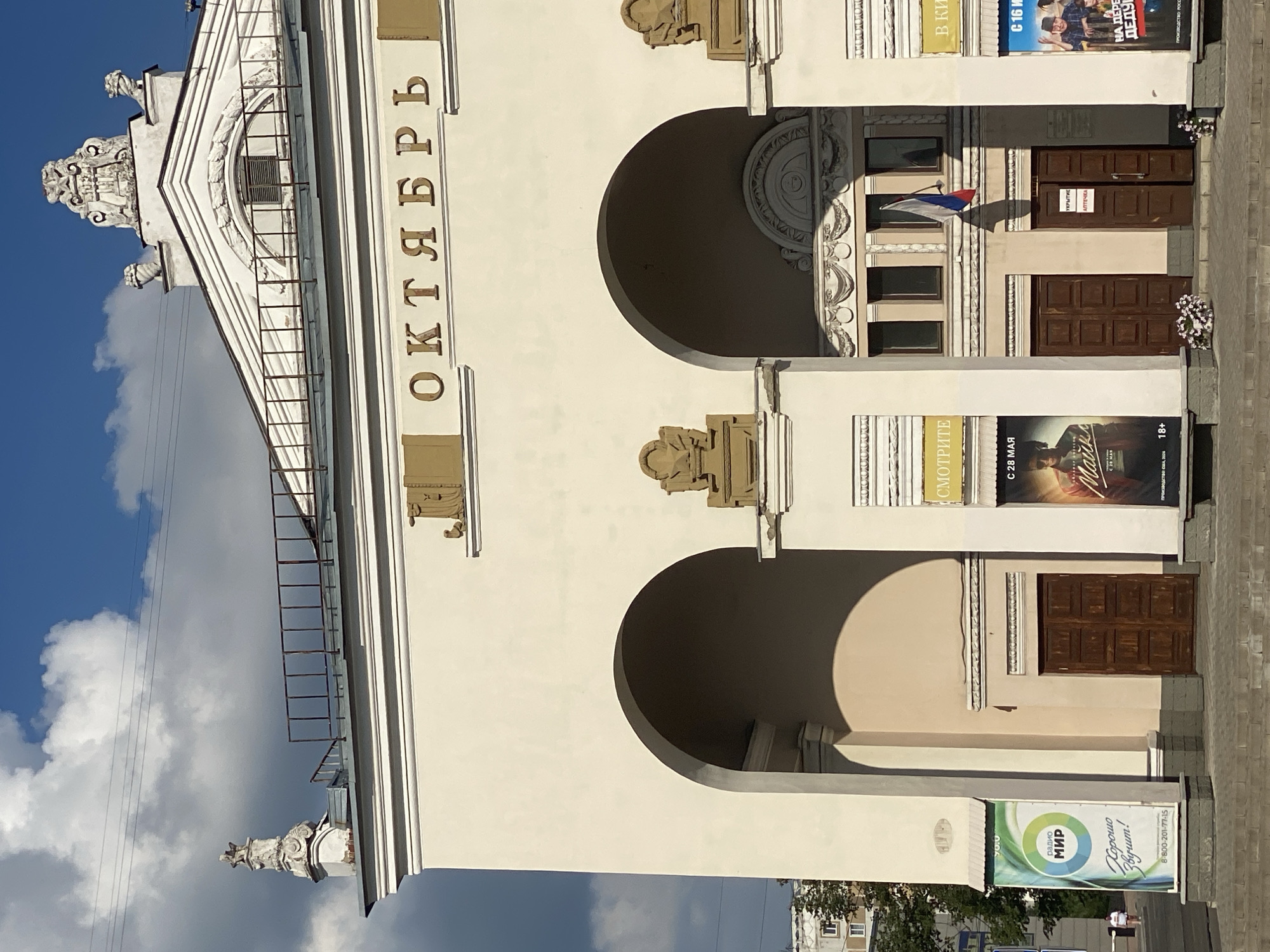 The image shows the facade of the "Oktyabr" (October) cinema in Novokuznetsk, Russia. The building features large arched entrances and is adorned with classical architectural elements, including decorative sculptures on the roofline and above the arches. The name of the cinema, "ОКТЯБРЬ," is prominently displayed in large, gilded letters on the upper facade. Several movie posters are visible on either side of the entrances, advertising films such as "Майор Гром" and "На последнем рубеже." A Russian flag is displayed above the central entrance. The sky is blue with scattered white clouds, suggesting a sunny day. The scene appears to be a public space, possibly a street or square in front of the cinema, with paving stones and a few people walking in the background.