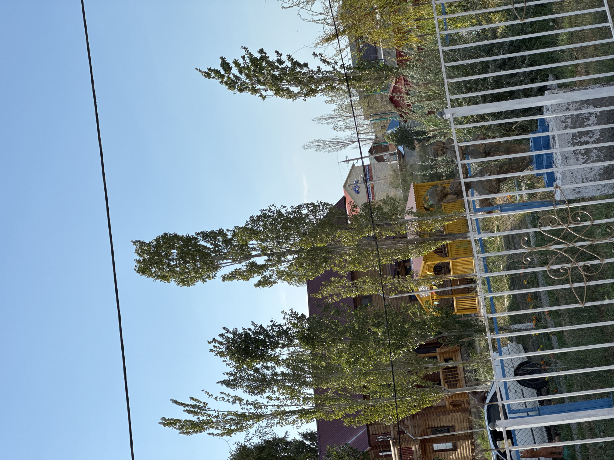 The image depicts a scene in the city of Zharbulak, Kazakhstan. In the foreground, a white metal fence with decorative scrollwork partially obscures the view. Behind the fence, a white car is parked on a grassy area. A small yellow gazebo with a brown roof is visible in the middle ground, situated between two tall trees with green foliage. To the left of the gazebo, a large wooden house with a reddish-brown roof is prominent. The house features balconies and multiple windows. Behind the wooden house and gazebo, various other buildings with different colored roofs and structures can be seen. Several tall trees with green leaves are scattered throughout the scene. The sky above is a clear, bright blue. The overall environment appears to be a residential or recreational area.