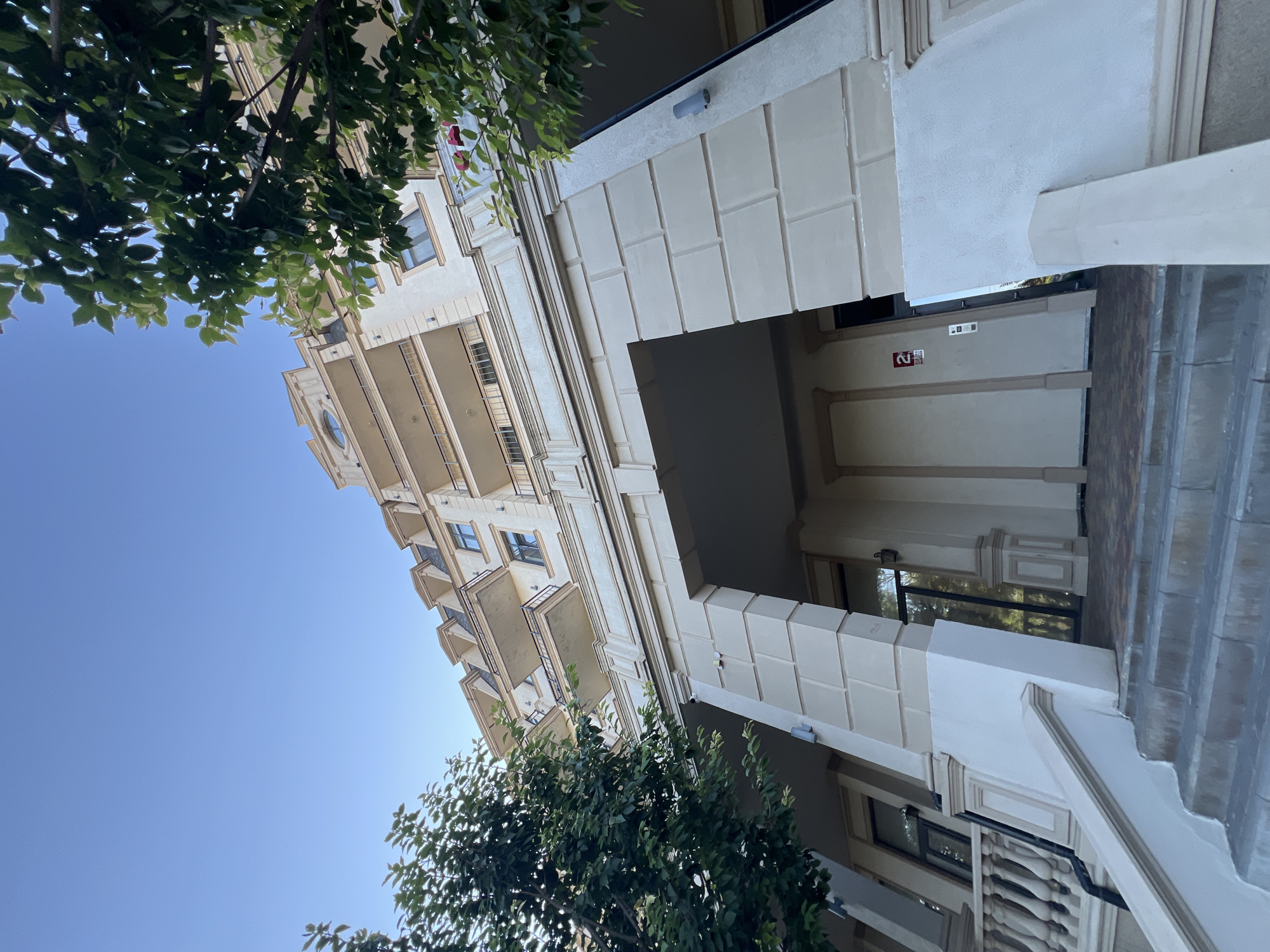 This is a low-angle, full-shot, eye-level, exterior photograph taken in Fontanka, Ukraine. The image captures the facade of a multi-story building with a clear blue sky above. The building features a light-colored, ornate exterior with multiple balconies, mansard roofs, and decorative molding. The entrance to the building is visible under an archway, with a staircase leading up to it. Trees with green foliage frame the building on both sides, partially obscuring the upper floors. A red sign with a white number "2" is mounted on the wall near the entrance, and an intercom panel is also visible. The overall scene suggests a residential or commercial building in a well-maintained urban environment.