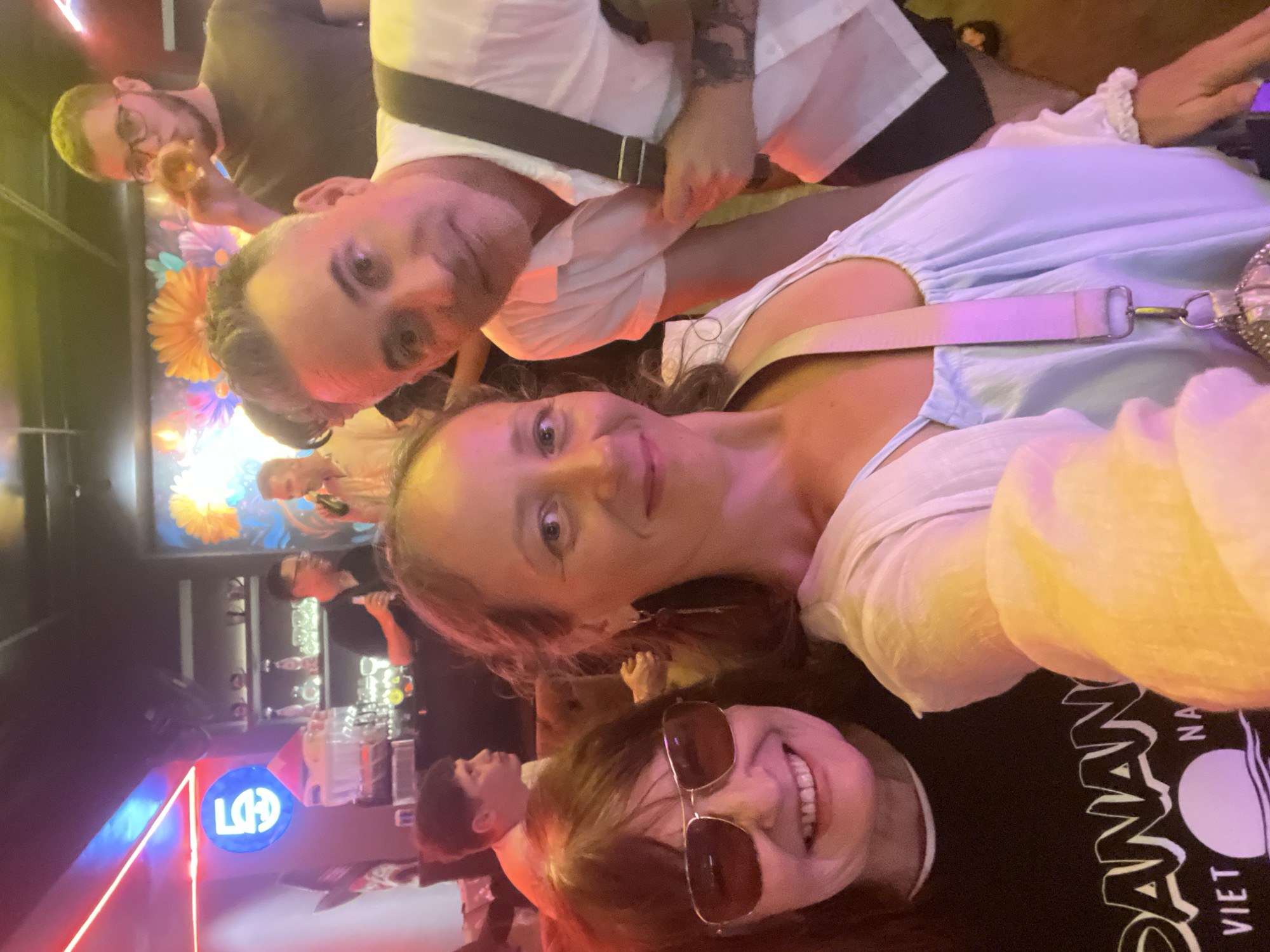 A group of five people are gathered in what appears to be a bar or nightclub. In the foreground, two women are taking a selfie, with one smiling broadly and wearing sunglasses, and the other looking towards the camera with a slight smile. Behind them, a man in a light-colored shirt is visible, along with another man in a dark shirt and glasses, who is holding a drink. Further in the background, another person is visible, partially obscured. The environment features neon lighting, shelving with bottles, and a colorful mural on the wall, suggesting a lively entertainment venue, possibly in the city of Đà Nẵng, Vietnam, given the text on one of the woman's shirts.