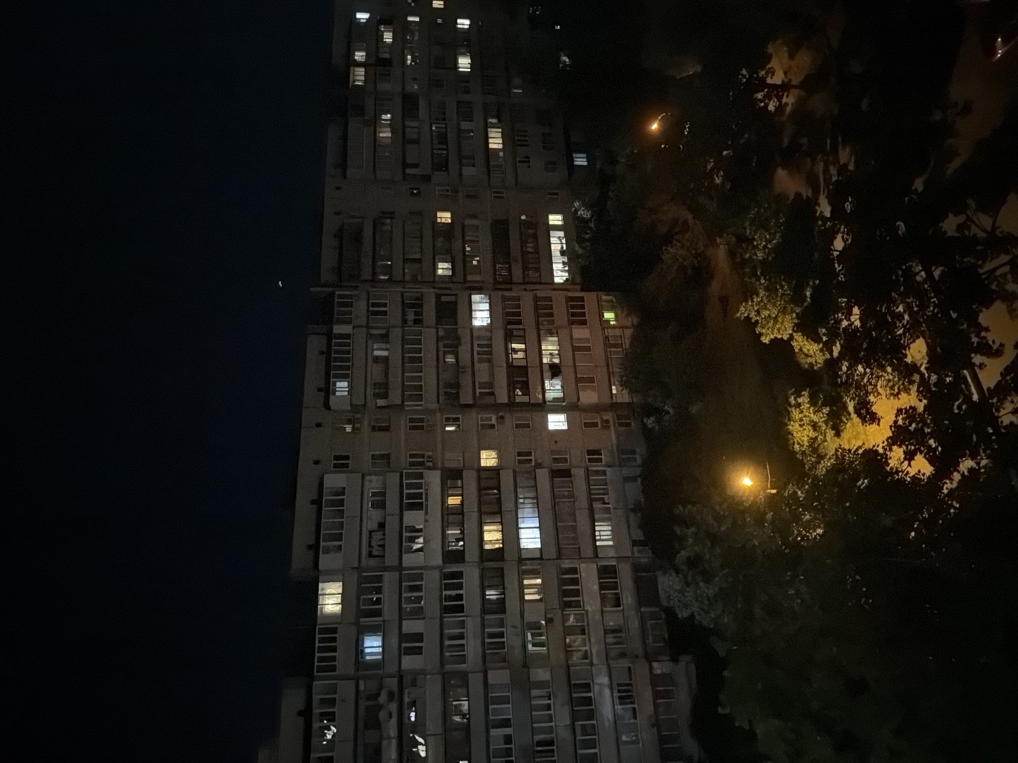 A nighttime aerial shot shows a multi-story apartment building with numerous lit windows. The building is long and appears to be made of concrete panels. Trees and foliage surround the base of the building, and a streetlamp illuminates a portion of the ground with a warm glow. The sky above is dark and appears clear, with a faint crescent moon visible. The overall scene depicts a residential area at night, with lights indicating ongoing activity within the apartments.