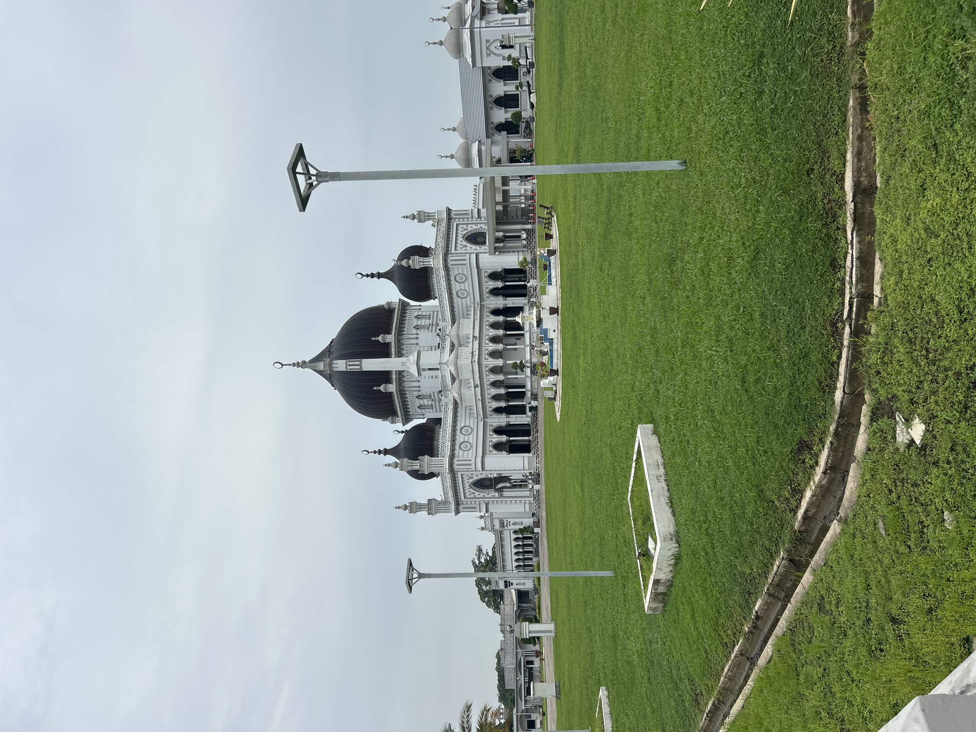 The image shows the exterior of Zahir Mosque in Alor Setar, Malaysia, on an overcast day. The mosque is a large, white building with multiple dark domes and minarets. The main dome is prominent and centrally located. A manicured green lawn surrounds the mosque, with several streetlights and some decorative landscaping visible. In the foreground, there is a drainage ditch and a concrete planter. The overall scene is serene and architectural.