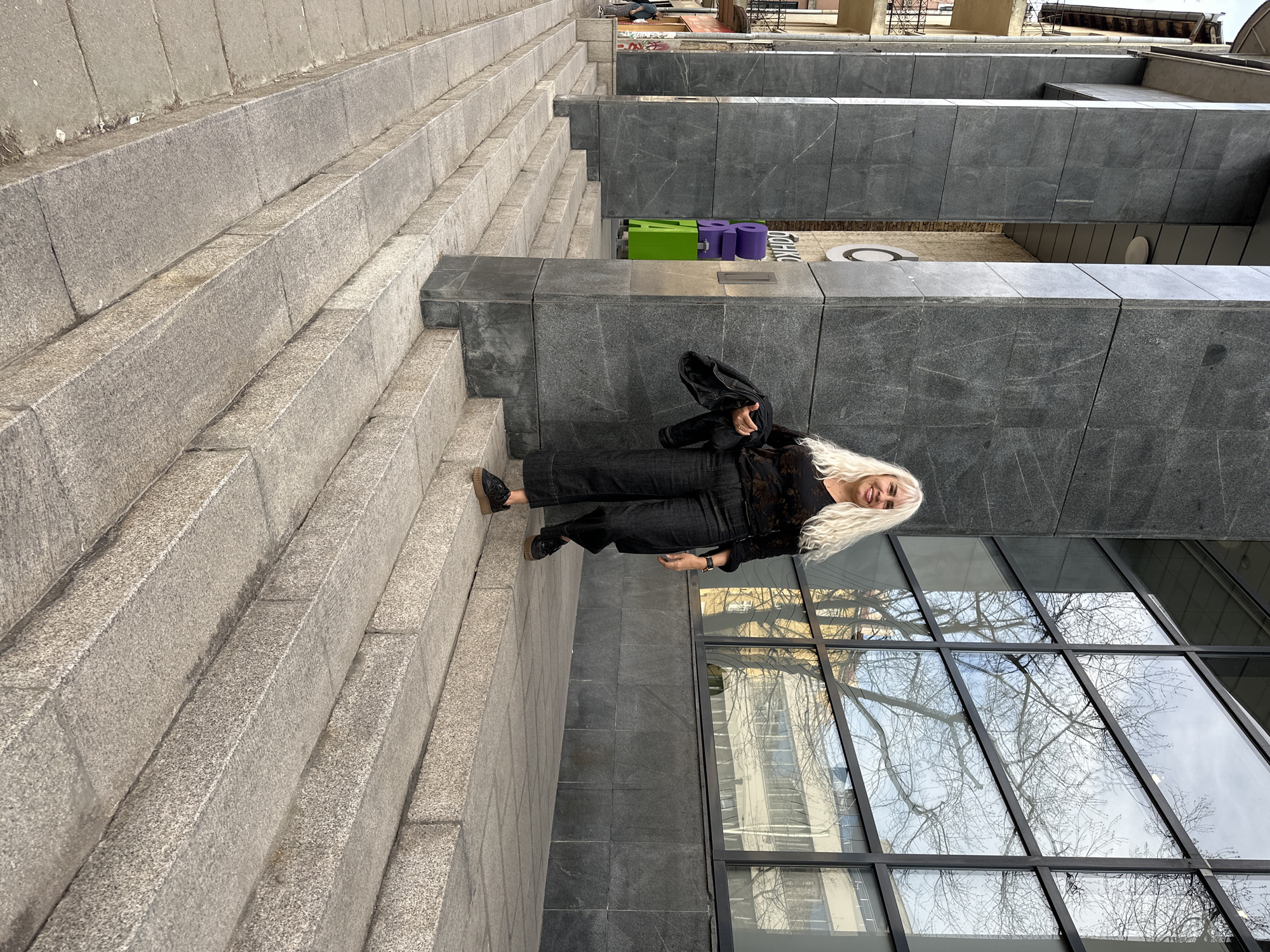 The image shows a woman with long blonde hair standing on stone steps next to a modern building with large windows. The building reflects the sky and bare tree branches. The steps appear to be part of a walkway leading up to the building. A green and purple object is visible further up the steps. The photo was taken outside of the National Palace of Culture in Sofia, Bulgaria.