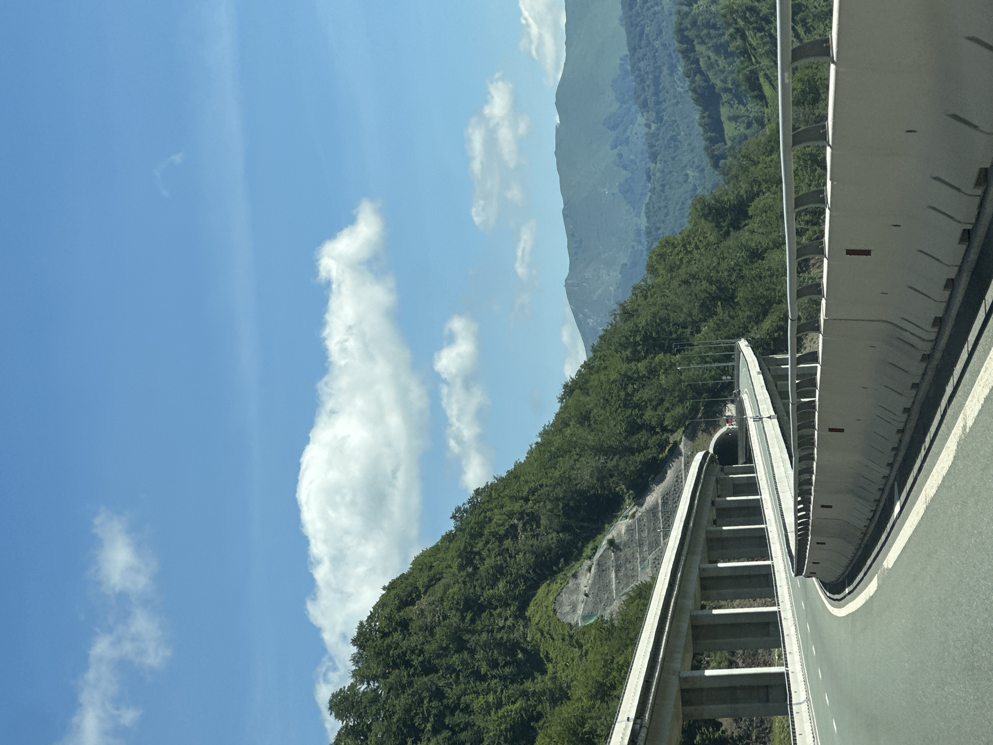 The image shows a winding highway cutting through a lush, green mountainous landscape. The highway itself is elevated in sections, supported by concrete pillars, and has concrete barriers along the side. A tunnel entrance is visible in the background, carved into the mountainside. The mountains are covered in dense forest, and the sky above is a clear blue with scattered white clouds. The scene appears to be a view from a moving vehicle on the highway. The specific location in Klopot, Montenegro, cannot be determined from the visual information provided in the image alone.