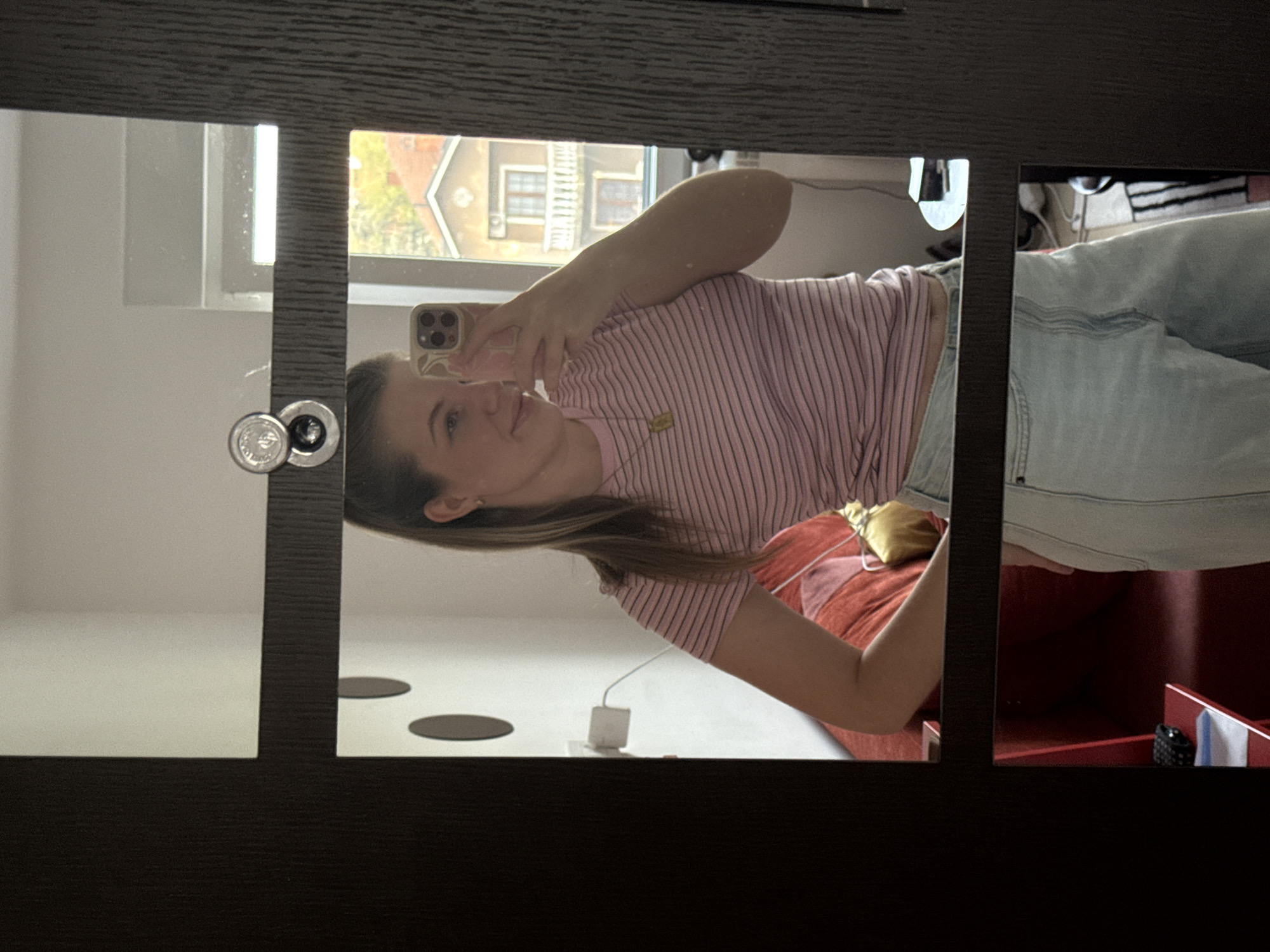 An indoor mirror selfie shows a young woman taking her picture. The image is rotated 90 degrees counter-clockwise. The woman, with long brown hair, is wearing a pink and white striped short-sleeved t-shirt and light blue jeans. She is holding an iPhone, which has a light-colored case and three camera lenses, to capture her reflection in what appears to be a multi-paneled mirror, possibly part of a wardrobe or door. She has a slight, calm smile.
The mirror's dark frame sections divide the view. Reflected in the upper right mirror panel, a window reveals residential buildings with sloped roofs and some foliage, suggesting an urban or suburban setting. This likely indicates it is daytime, possibly late afternoon, due to the natural light.
Behind the woman, a bed or sofa with red and pink patterned bedding and a yellow decorative pillow is visible. On a surface below the mirror, a white charging block with a cable is plugged in, and two dark circular objects are present. In the lower right mirror section, a dark patterned bag and a folded white and blue cloth are visible inside a reddish piece of furniture, possibly a drawer or shelf. A silver knob or handle is also visible on the mirror frame. No discernible text is visible.