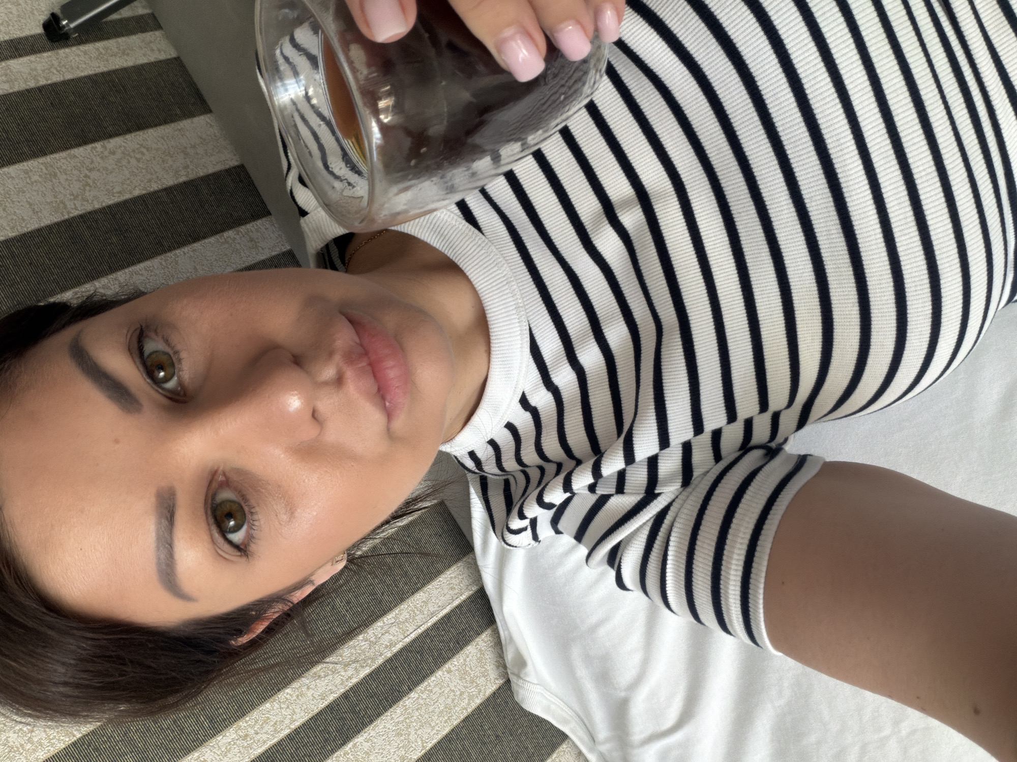 A woman with dark brown hair and hazel eyes is lying down and holding a glass of dark liquid. She is wearing a black and white striped shirt and a white shirt underneath. The background consists of striped fabric. The image is a selfie taken from a high angle. No specific location in Sochi, Russia is discernible from the image.