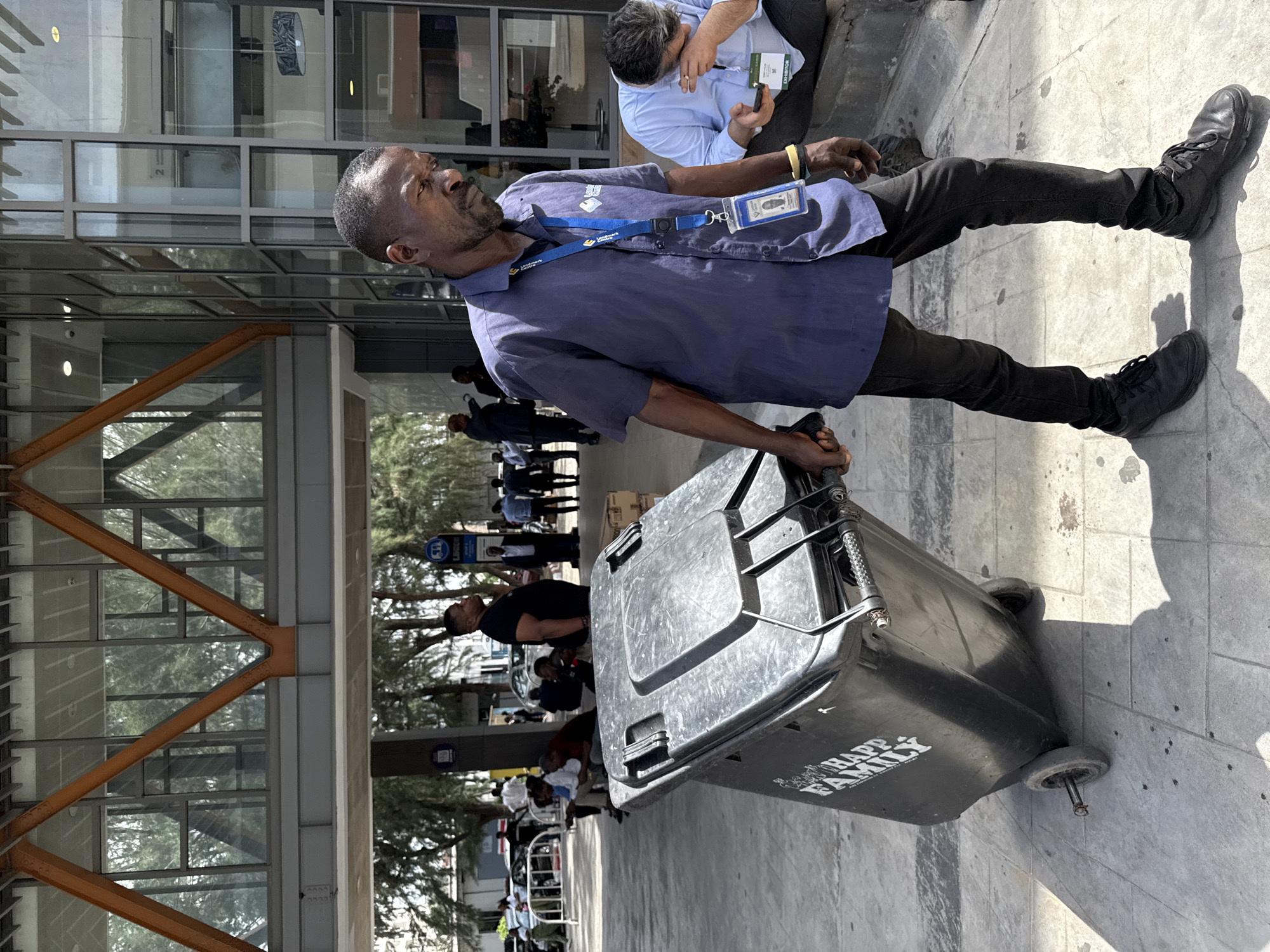 A man pulls a gray, wheeled trash can with "HAPPY FAMILY" printed on its side. He wears a blue shirt, black pants, black shoes, and a lanyard with a badge. He looks up and to his left. In the background, people are visible under the covered entrance of a building with glass windows. A man kneels on the right, looking at a small device. This photo was taken in Alaguntan, Nigeria.