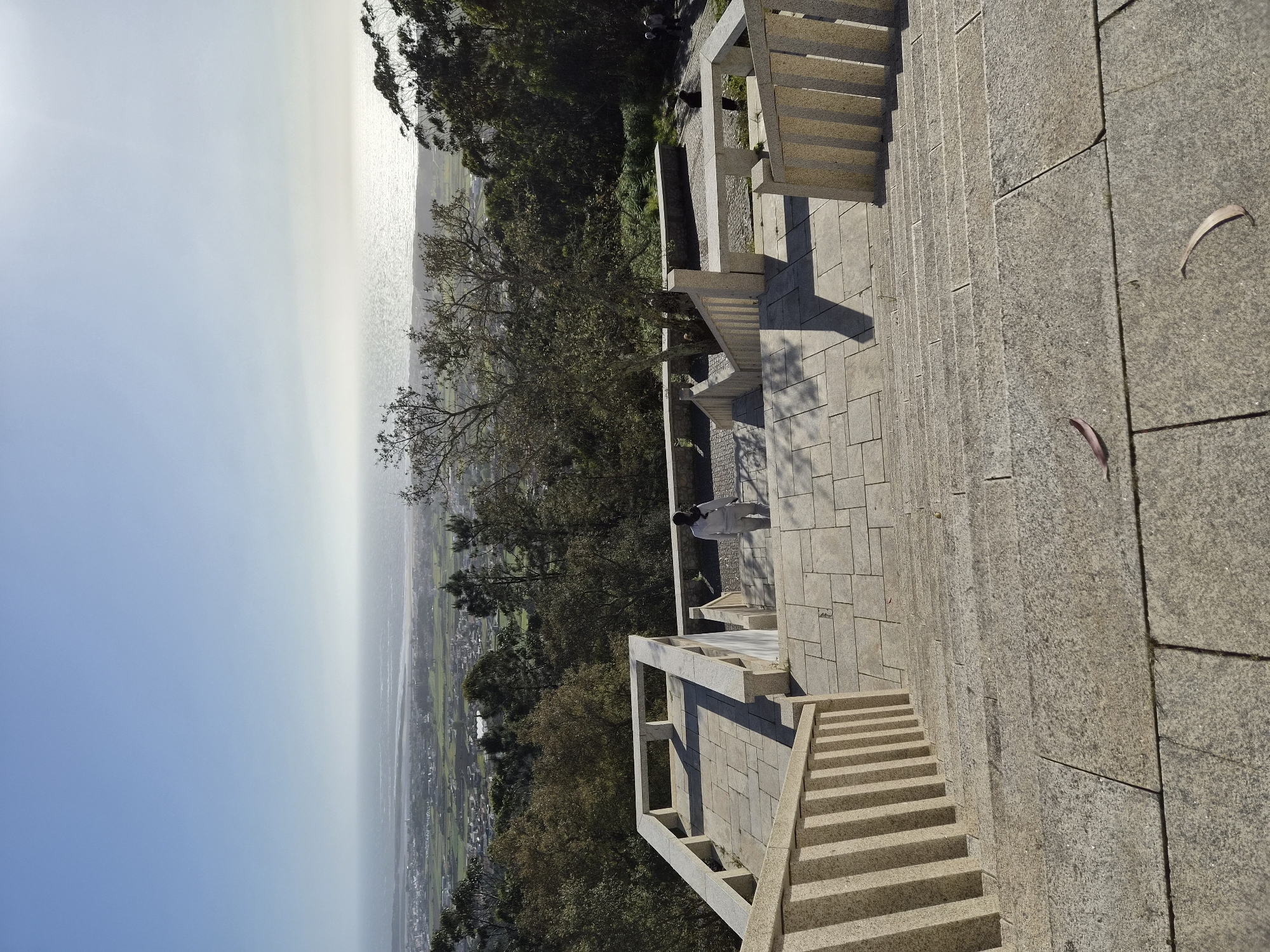 A high-angle shot captures an individual on a stone terrace in Esposende, Portugal, set against a backdrop of the Atlantic Ocean. The terrace is constructed of light-colored stone pavers and includes a series of steps. A person is visible on the terrace, dressed in white clothing. Beyond the terrace, the terrain slopes down, featuring trees and greenery. In the background, the Atlantic Ocean meets the horizon under a bright sky with the sun visible. The image suggests a scenic overlook point.